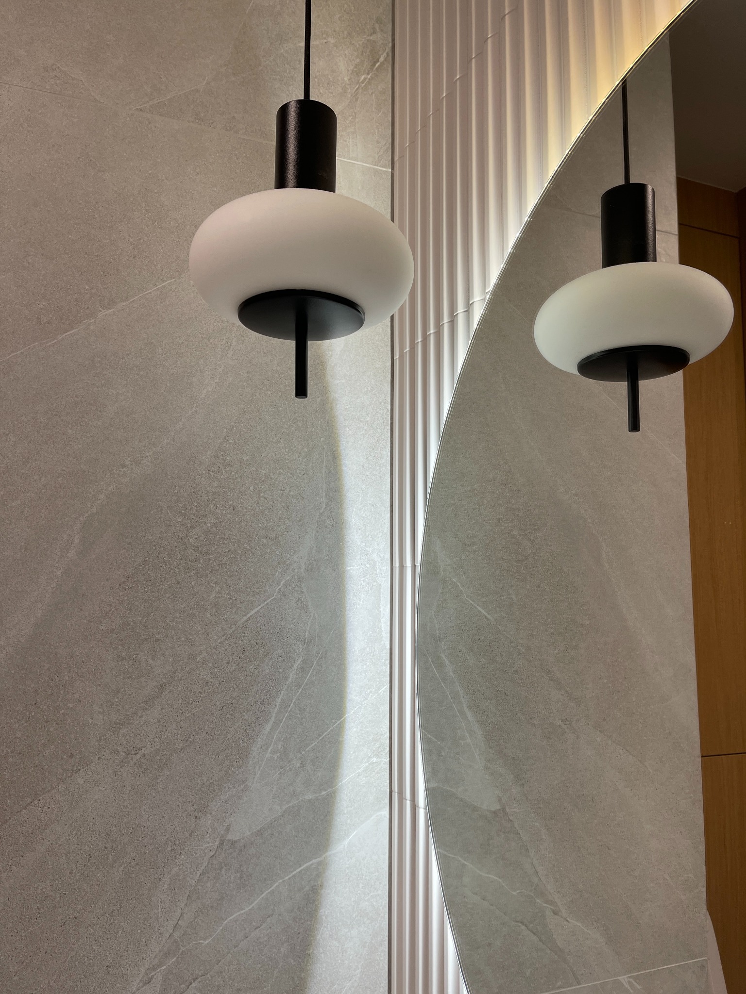 Szara ściana łazienki wykończona płytkami imitującymi kamień, z okrągłym lustrem i nowoczesną lampą wiszącą z czarnym elementem i mlecznym kloszem.