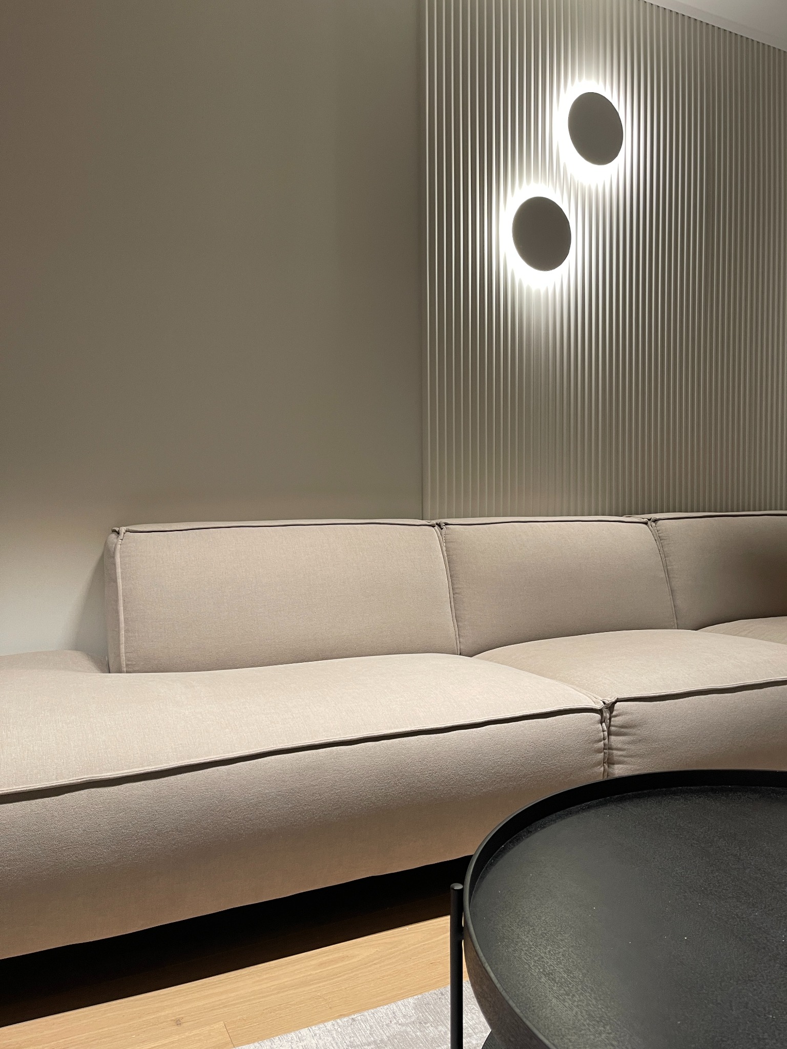 Fragment salonu z beżową sofą modułową, okrągłym czarnym stolikiem kawowym i ścianą z pionowymi panelami oraz dwoma okrągłymi lampami LED.