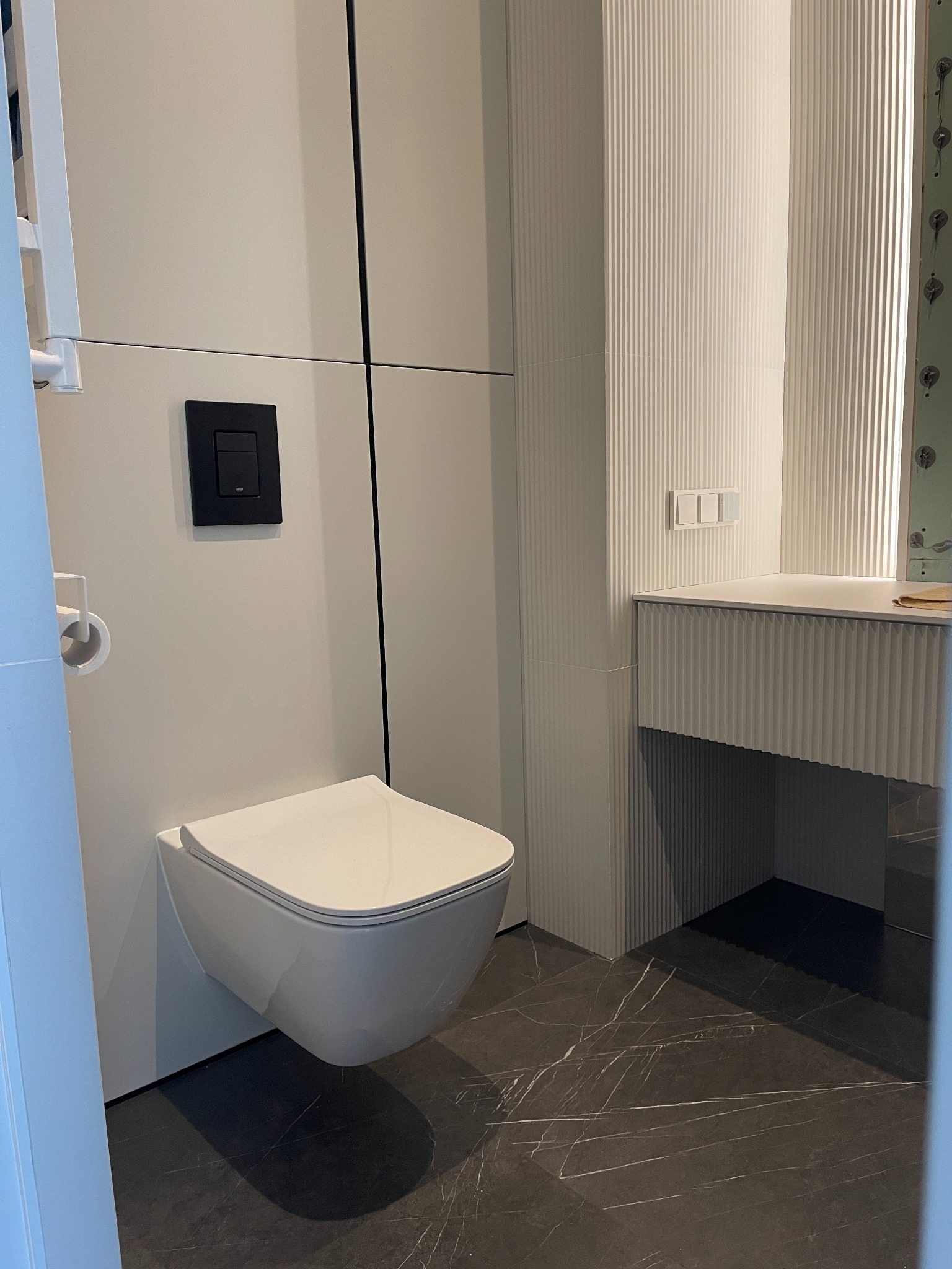 Nowoczesna łazienka z podwieszaną toaletą, czarnym przyciskiem spłuczki i marmurową podłogą, ściany wykończone w białych, pionowych panelach.