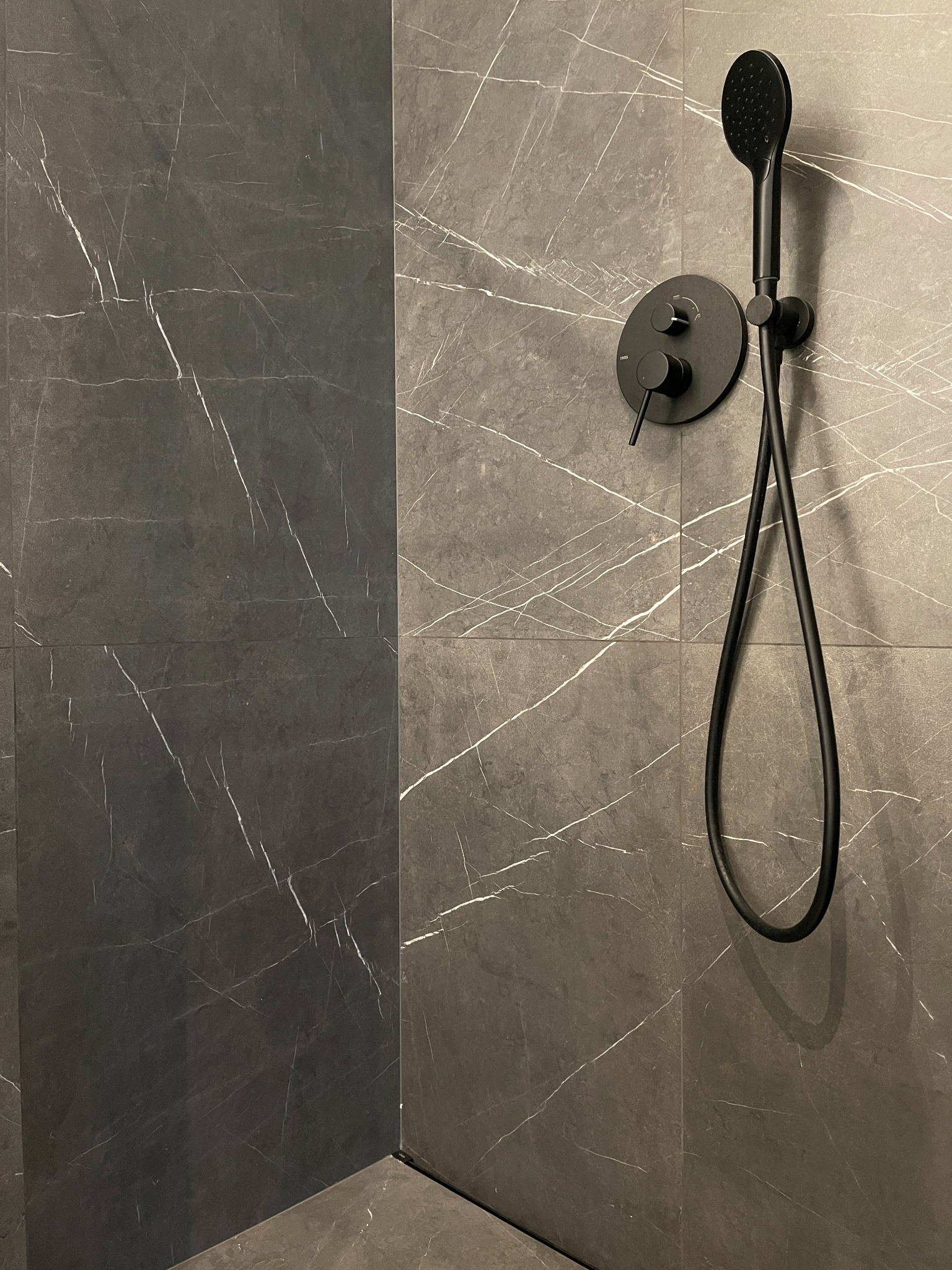 Nowoczesna kabina prysznicowa z ciemnoszarymi płytkami imitującymi marmur i czarną armaturą, w tym deszczownicą i baterią.
