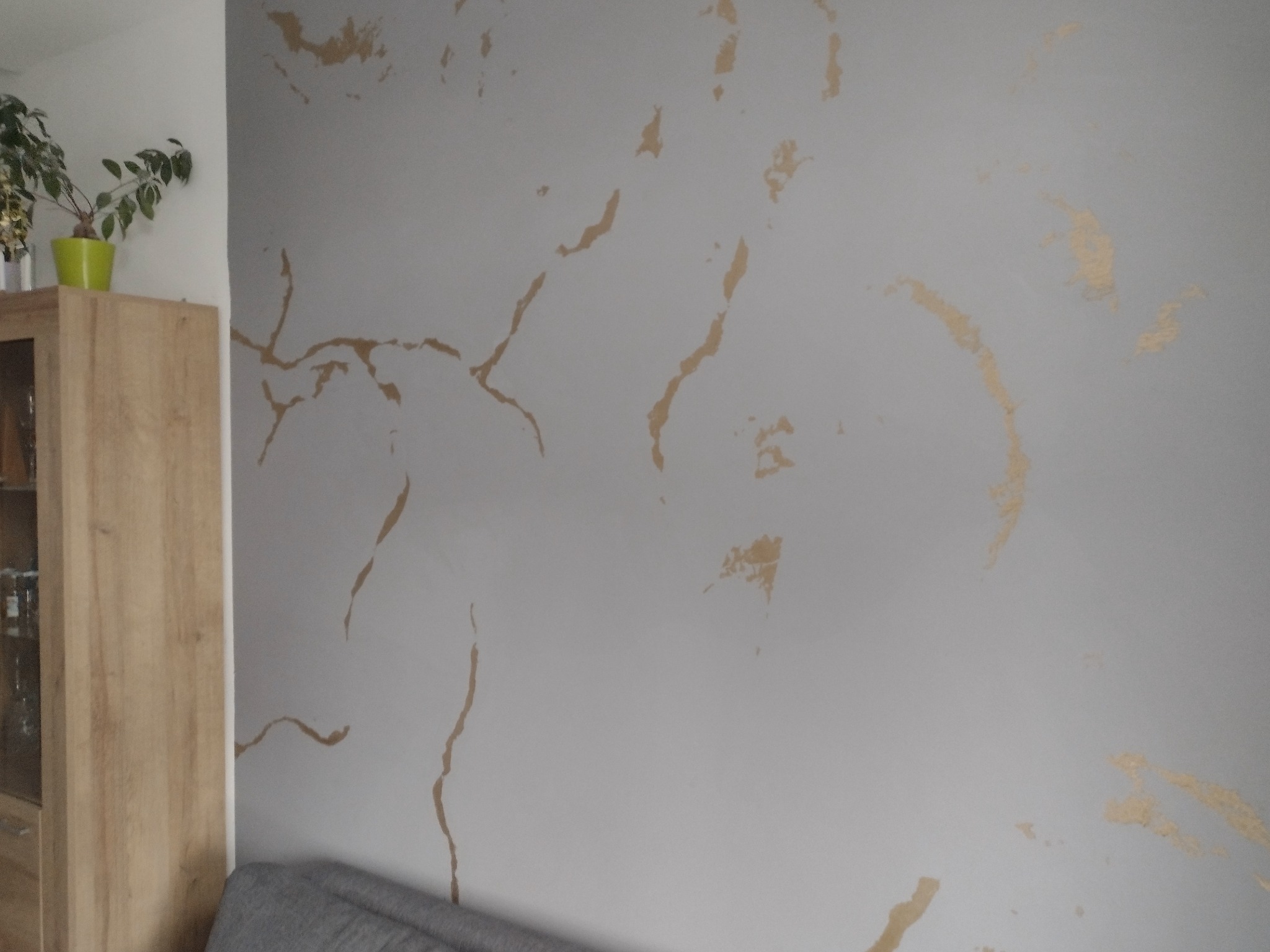 Szara ściana z dekoracyjnym wzorem imitującym marmur w odcieniach złota, widoczny fragment drewnianej szafki z rośliną w zielonej doniczce oraz fragment szarej sofy.