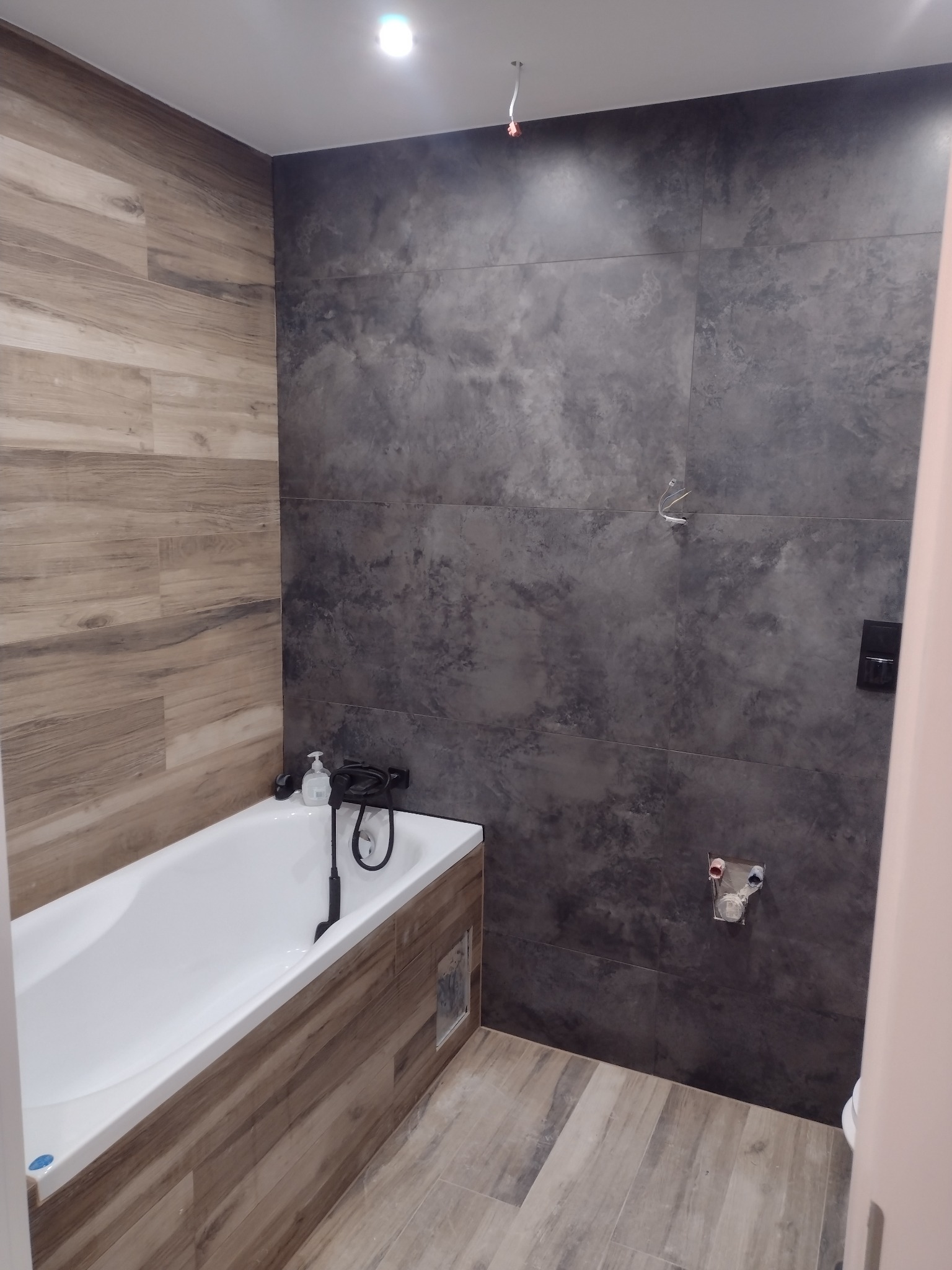 Wykończona łazienka z wanną obudowaną panelami imitującymi drewno, ciemnoszare płytki imitujące beton na ścianie, widoczne przyłącza wodne i wystające przewody elektryczne.
