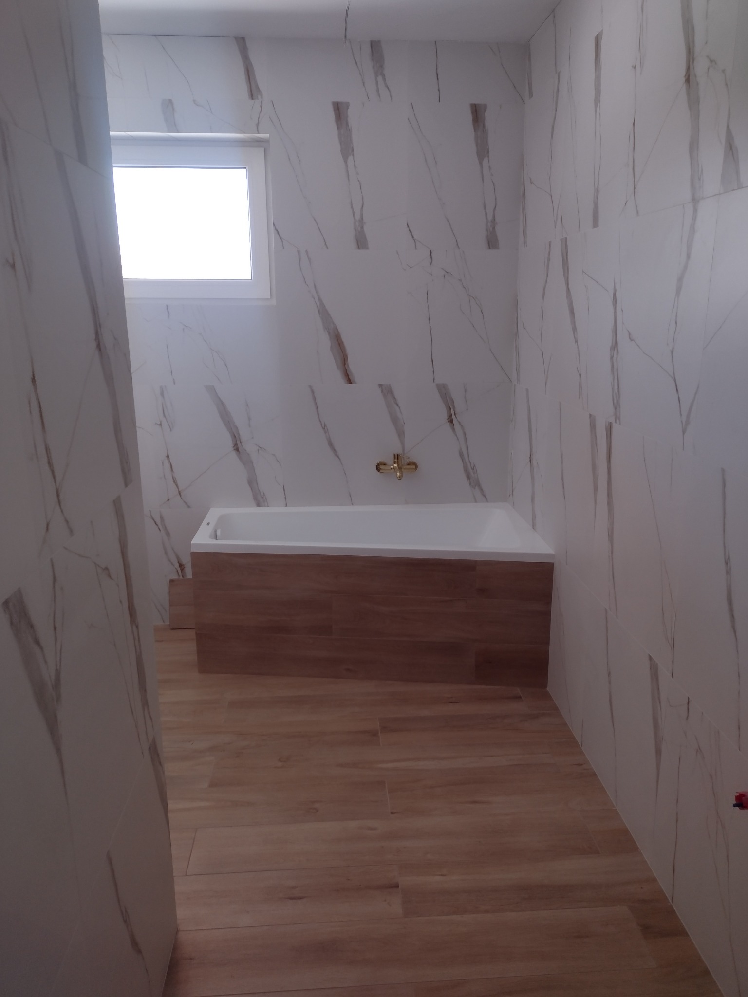 Wykończona łazienka z wanną obudowaną drewnopodobną płytą, ściany wyłożone płytkami imitującymi marmur z brązowymi żyłami, drewniana podłoga i złota bateria. Okno z białymi ramami w górnym lewym rogu.