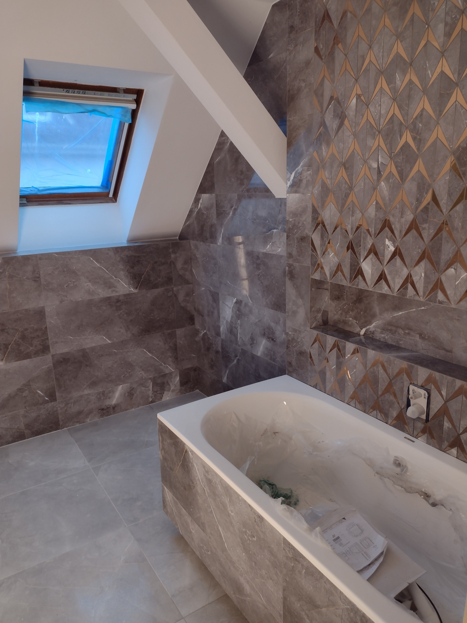 Wnętrze łazienki w trakcie remontu na poddaszu: szare płytki marmurowe z geometrycznym wzorem na ścianach, biała wanna zabezpieczona folią, okno dachowe z niebieską folią ochronną.