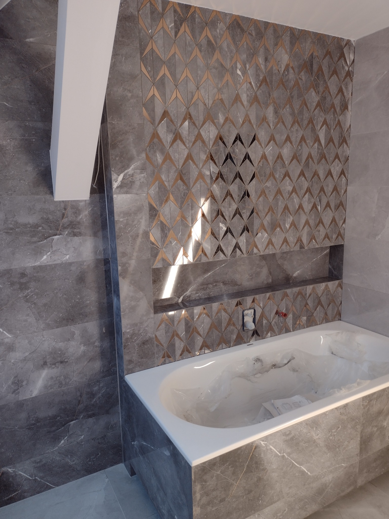 Wykończona łazienka z wanną obudowaną szarymi płytkami imitującymi marmur i ścianą z dekoracyjnymi płytkami w geometryczny wzór z akcentami w kolorze złota, wnęka w ścianie wykończona tymi samymi...