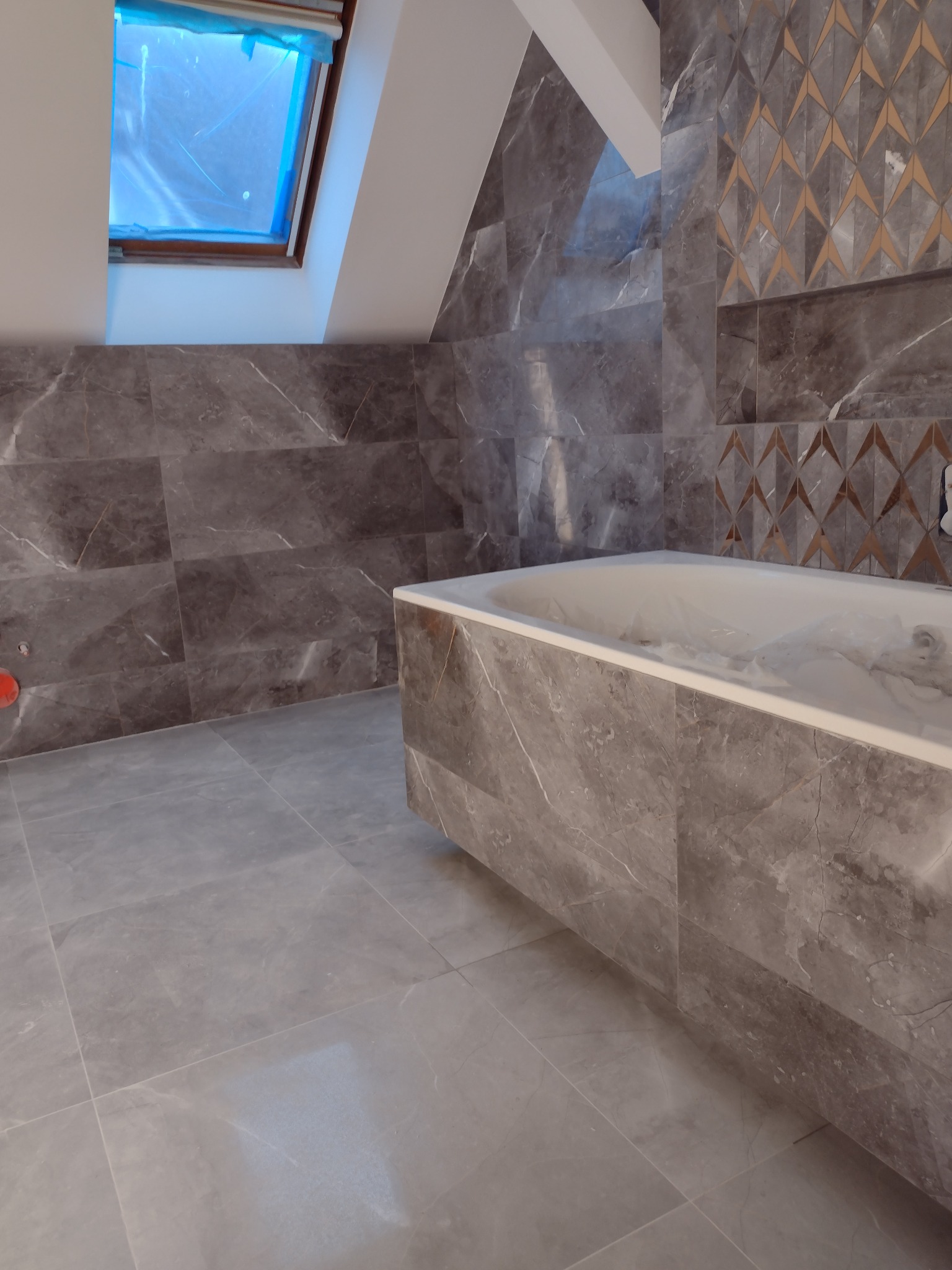 Wykończona łazienka na poddaszu z szarymi płytkami imitującymi marmur, dekoracyjnym pasem płytek z geometrycznym wzorem i wanną obłożoną tymi samymi płytkami; okno dachowe zabezpieczone folią...