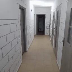 Korytarz z pomalowanymi ścianami imitującymi cegły, szare drzwi, beżowa podłoga z płytek.