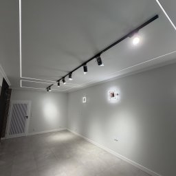 Nowoczesne wnętrze z szarą podłogą, białymi ścianami i designerskim oświetleniem sufitowym. Widoczne listwy LED i reflektory na szynoprzewodzie.