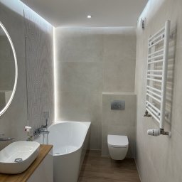 Nowoczesna łazienka z drewnopodobną podłogą, owalną umywalką na drewnianym blacie, białą wanną i grzejnikiem drabinkowym. Subtelne oświetlenie LED.