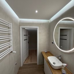 Nowoczesna łazienka po remoncie: beżowe płytki, okrągłe lustro z podświetleniem LED, drewniany blat z białą umywalką, grzejnik drabinkowy i otwarte drzwi.