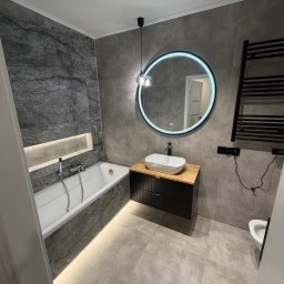 JANBUD - Nowoczesna łazienka w szarościach z podświetlaną wanną, okrągłym lustrem LED i drewnianą szafką pod umywalką. Minimalistyczny design z czarnym grzejnikiem.