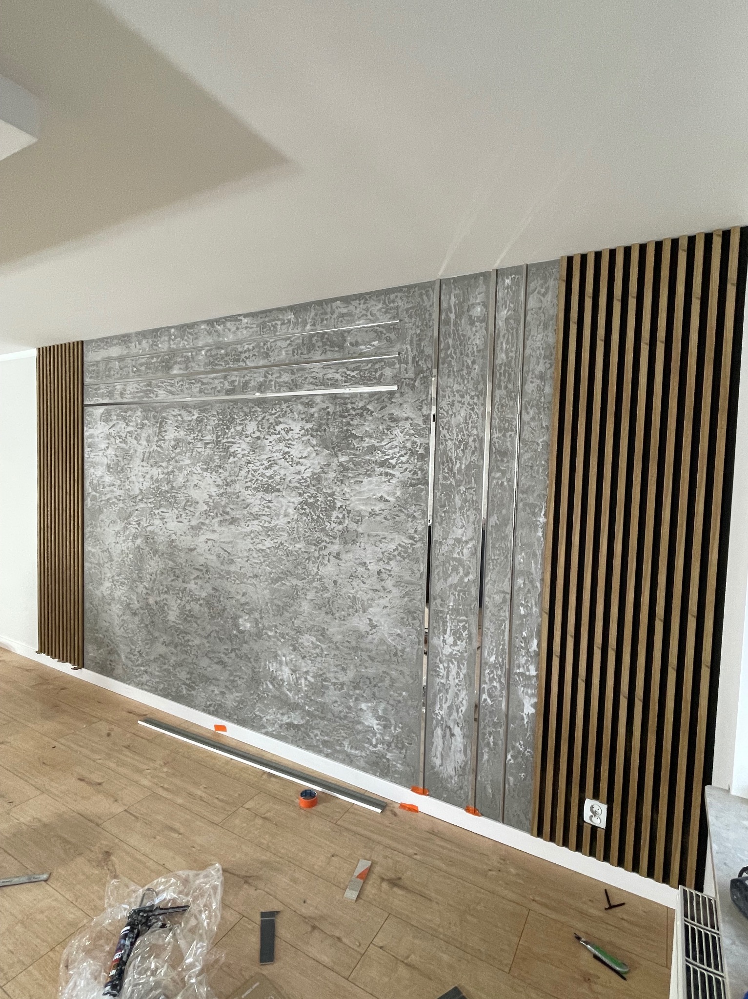 Wykończona ściana z dekoracyjnym tynkiem strukturalnym, drewnianymi lamelami i metalowymi listwami. Widoczne narzędzia i akcesoria montażowe na podłodze.