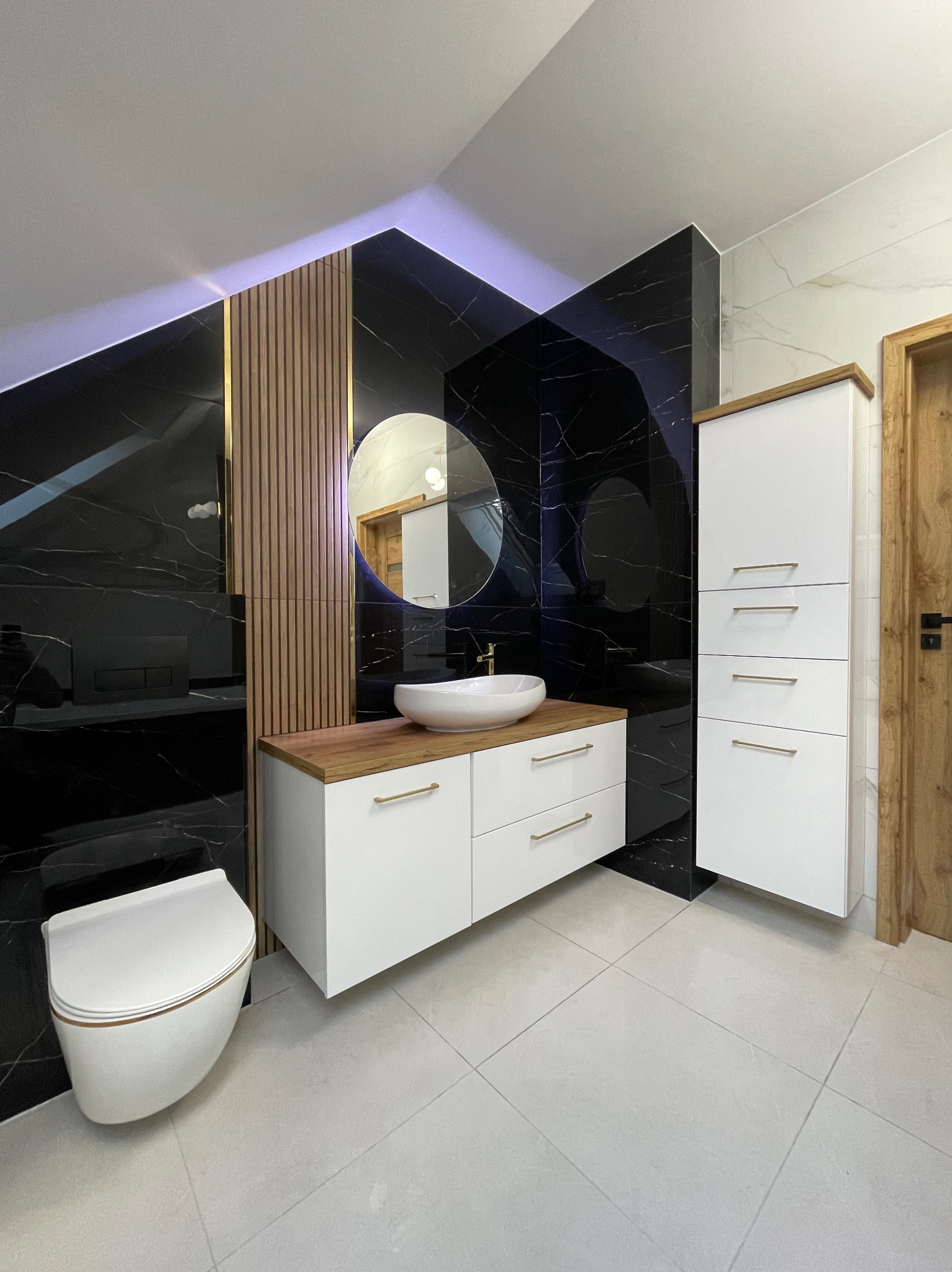 Nowoczesna łazienka z czarnymi płytkami imitującymi marmur, białymi szafkami z drewnianymi blatami, okrągłym lustrem i podświetleniem LED. Uwagę zwraca minimalistyczna toaleta.