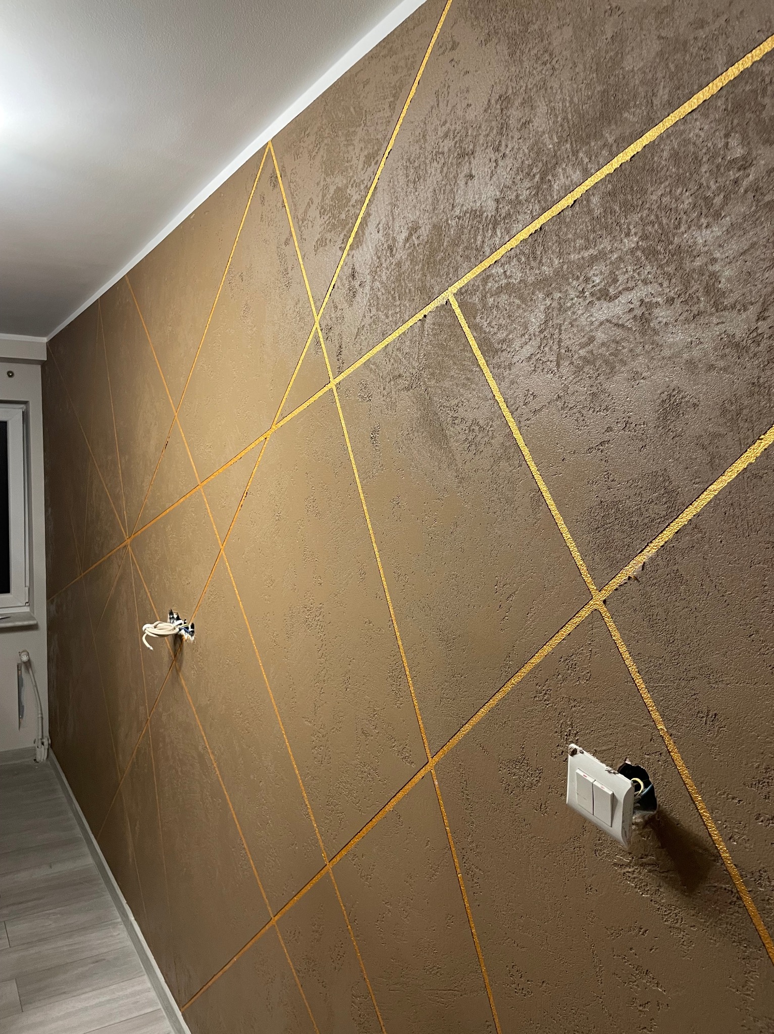 Dekoracyjna ściana w odcieniach brązu z geometrycznym wzorem ze złotych linii. Widoczne gniazdka elektryczne w trakcie montażu. Wykończenie wnętrza w nowoczesnym stylu.