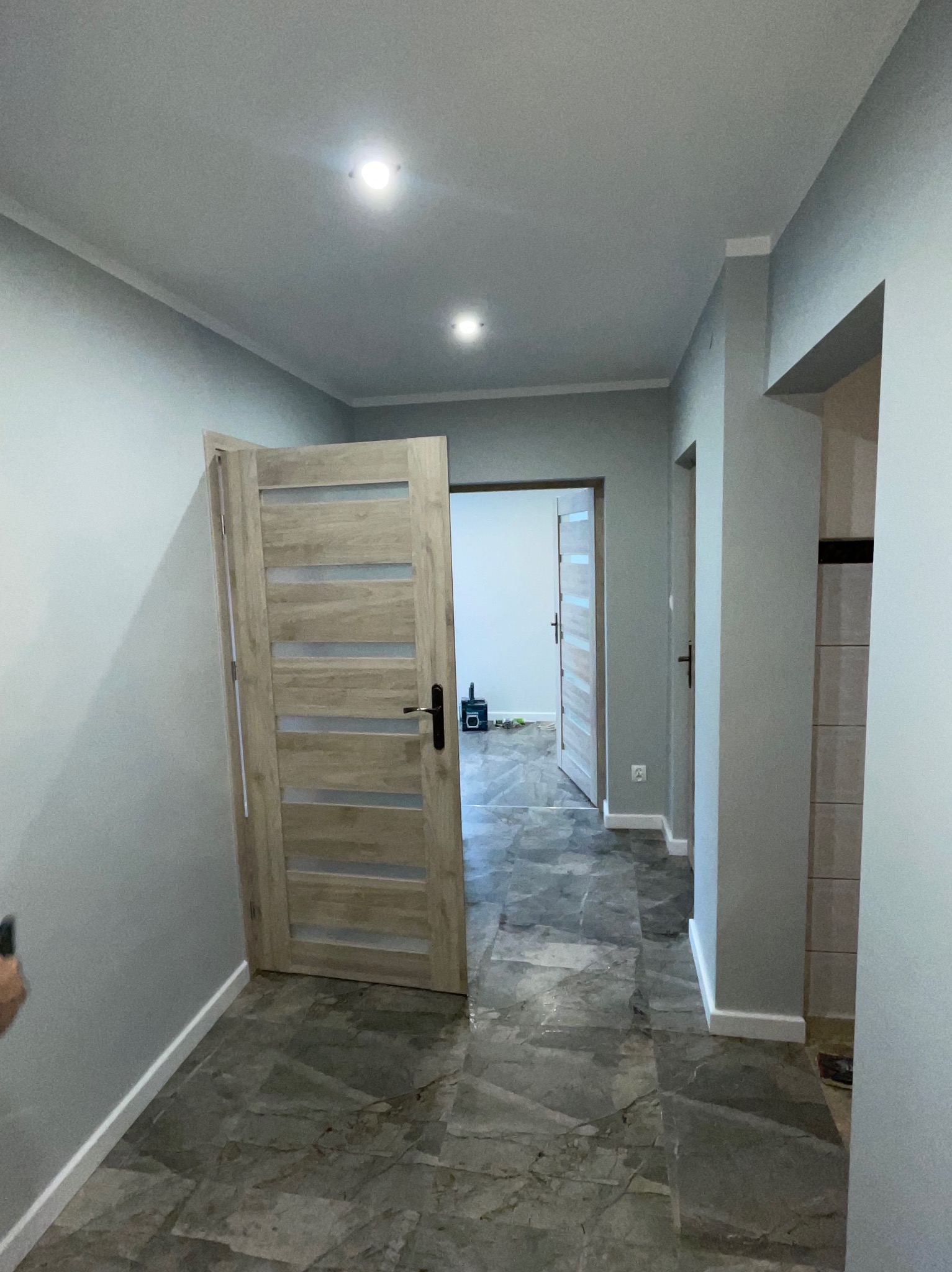 Jasne, nowoczesne wnętrze z szarą marmurową podłogą i drewnianymi drzwiami z przeszkleniami. Widoczne listwy przypodłogowe i oświetlenie sufitowe.