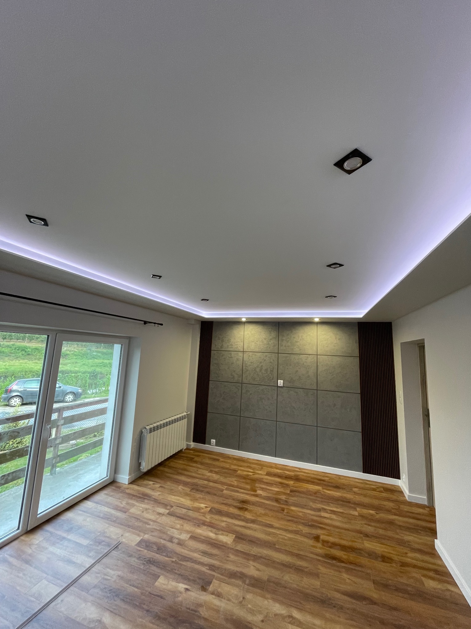 Nowoczesne wnętrze z panelami ściennymi, drewnianą podłogą i oświetleniem LED. Widok na balkon przez duże okno.