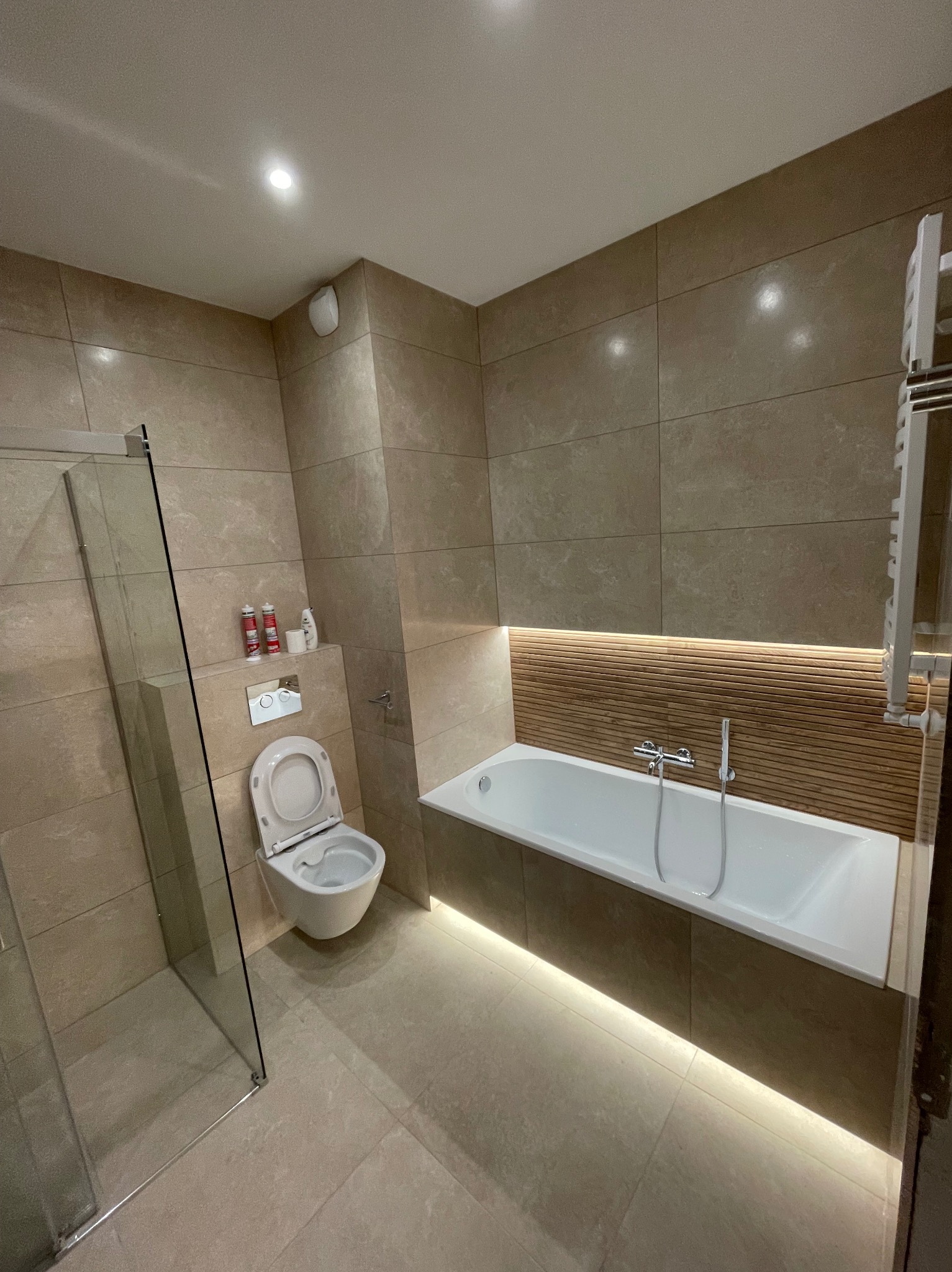Beżowa łazienka po remoncie z kabiną prysznicową, toaletą, wanną z podświetleniem LED i grzejnikiem drabinkowym. Użyto płytek imitujących kamień.