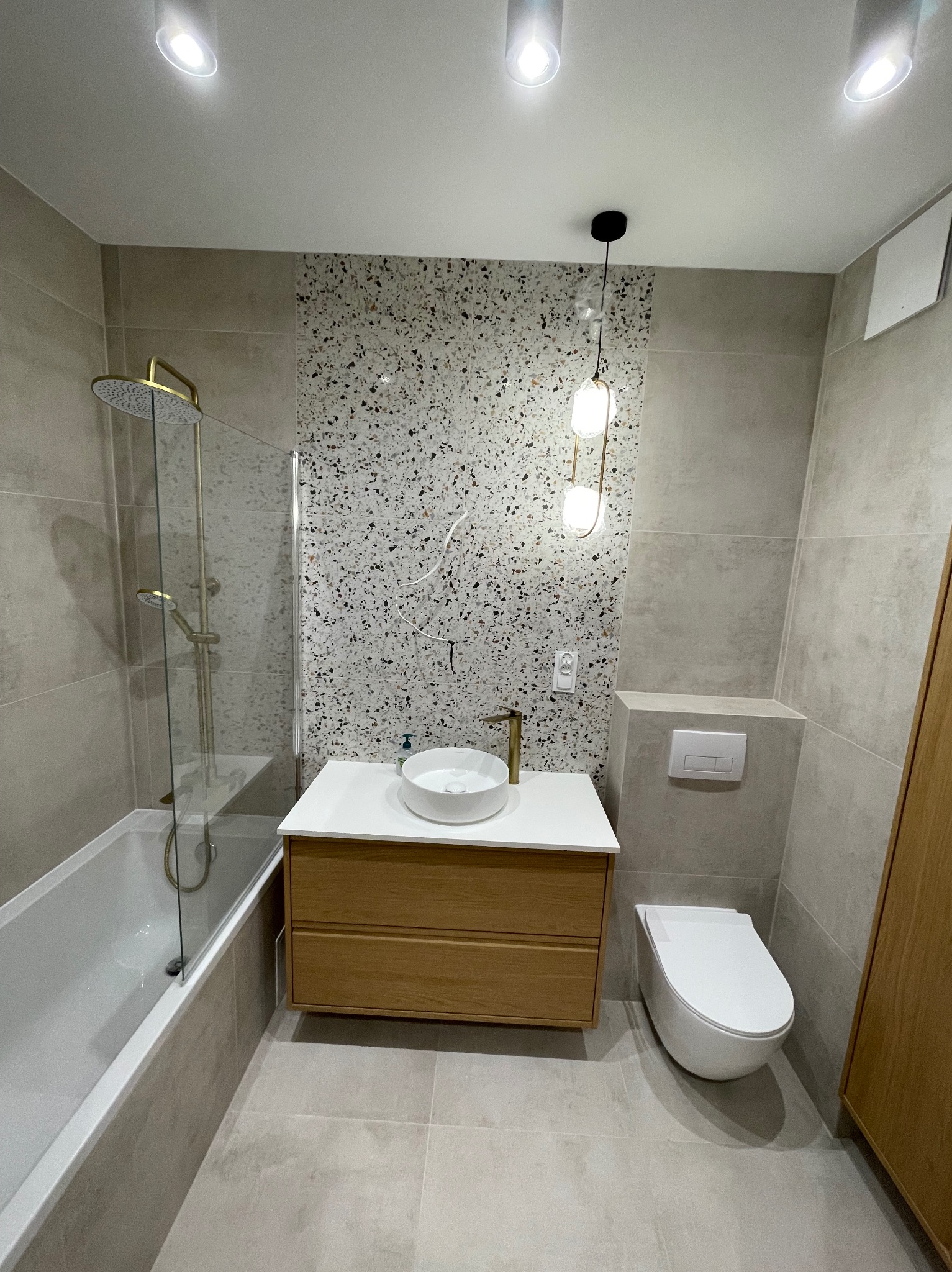 Nowoczesna łazienka w beżach z wanną, umywalką na szafce z jasnego drewna i toaletą. Ściana wykończona mozaiką. Oświetlenie punktowe i lampa wisząca.