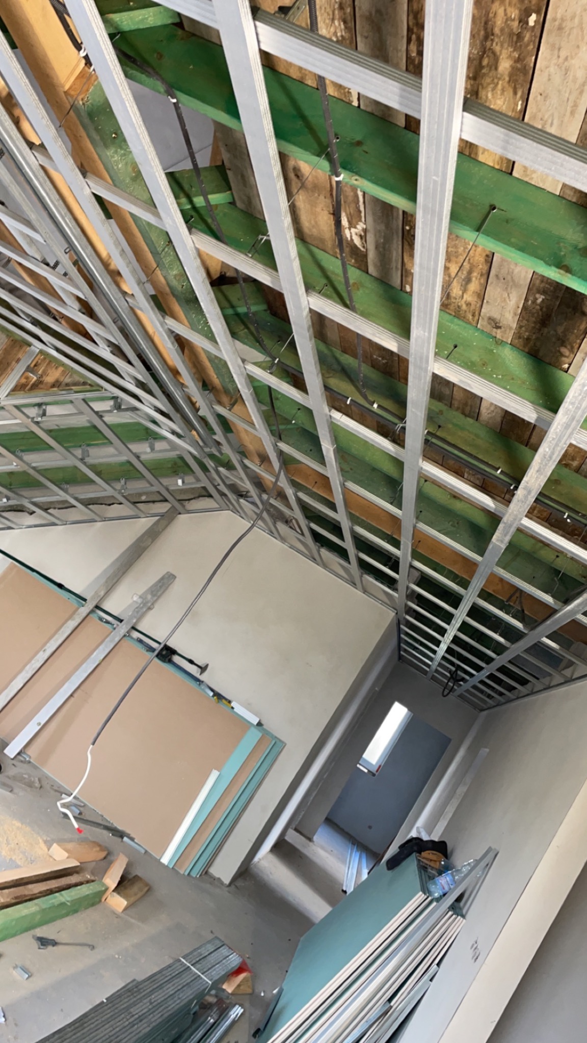 Szkielet sufitu podwieszanego z metalowych profili, widok z dołu, płyty gipsowo-kartonowe przygotowane do montażu, drewniane elementy konstrukcyjne w tle.