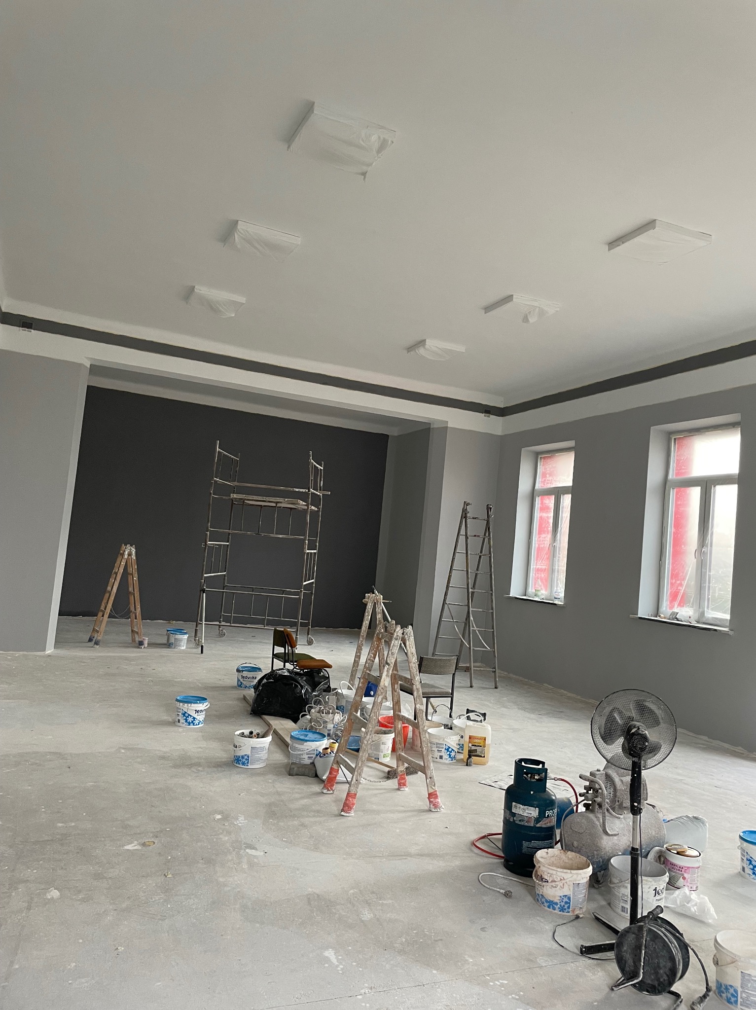 Pokój w trakcie malowania: szare ściany, biały sufit, drabiny, rusztowanie, pędzle, farby, wentylator. Lampy zabezpieczone folią. Widoczne przygotowania do malowania.