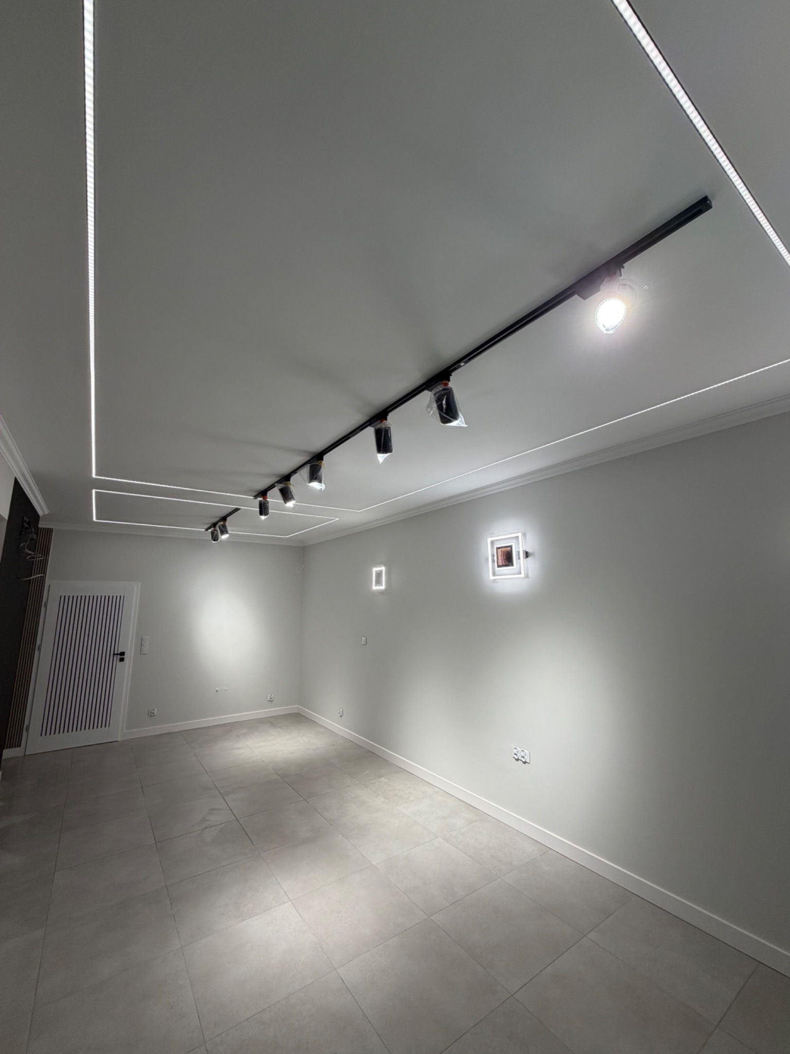 Nowoczesne wnętrze z szarą podłogą, białymi ścianami i designerskim oświetleniem sufitowym. Widoczne listwy LED i reflektory na szynoprzewodzie.