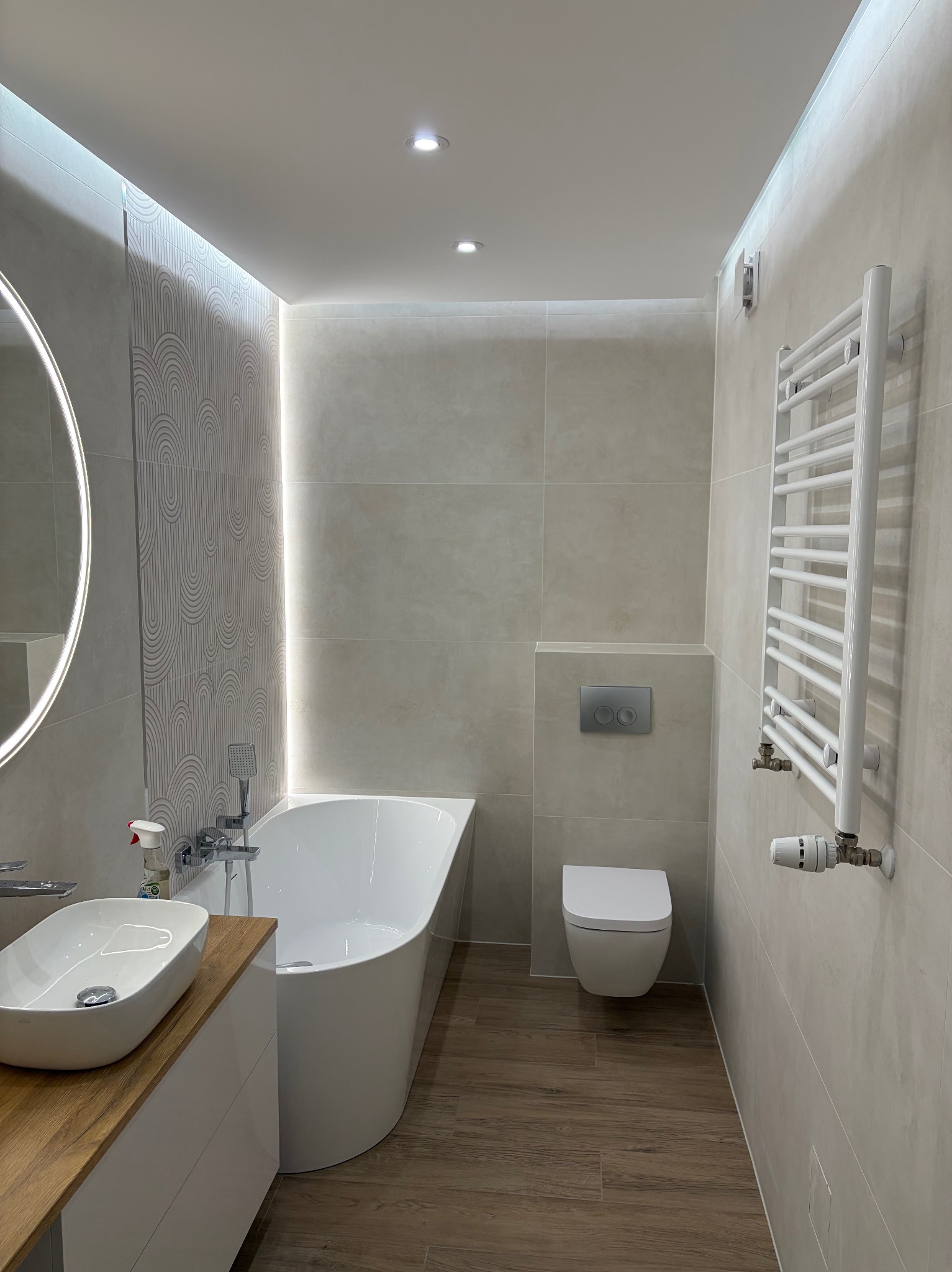 Nowoczesna łazienka z drewnopodobną podłogą, owalną umywalką na drewnianym blacie, białą wanną i grzejnikiem drabinkowym. Subtelne oświetlenie LED.