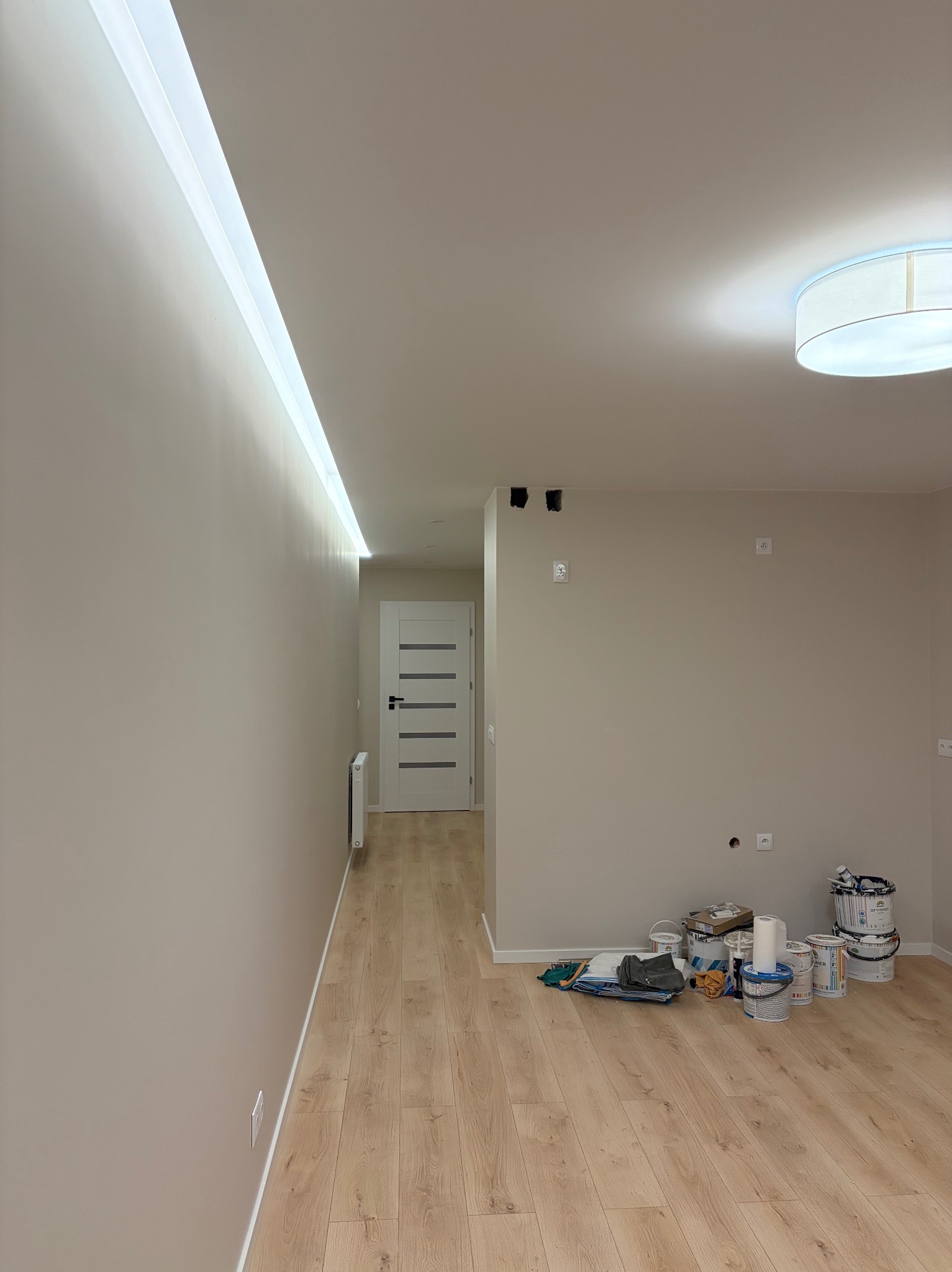 Jasne wnętrze po malowaniu: beżowe ściany z oświetleniem LED, drewniana podłoga, białe drzwi i farby malarskie. Widoczne gniazdka elektryczne i lampa sufitowa.