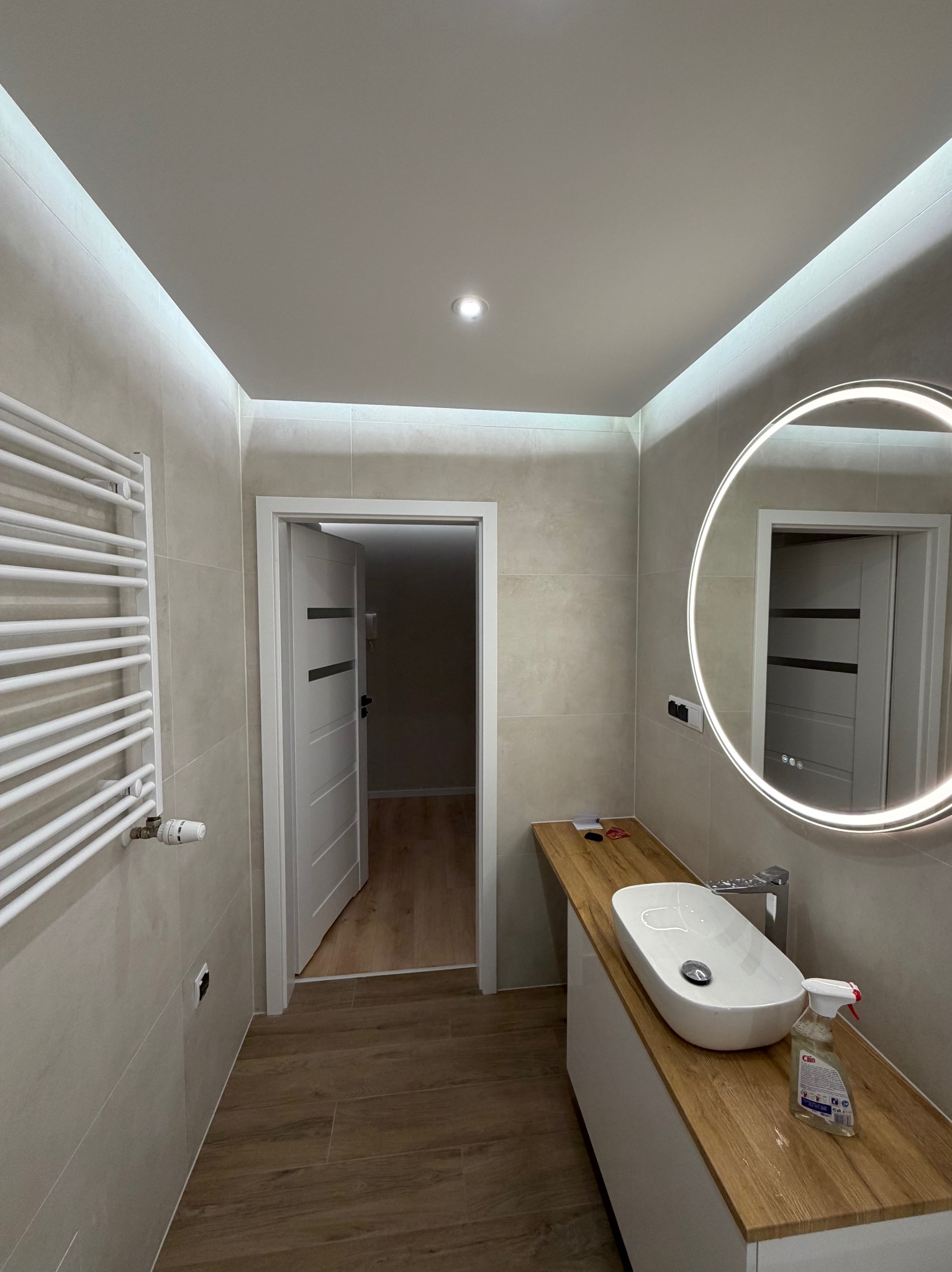 Nowoczesna łazienka po remoncie: beżowe płytki, okrągłe lustro z podświetleniem LED, drewniany blat z białą umywalką, grzejnik drabinkowy i otwarte drzwi.