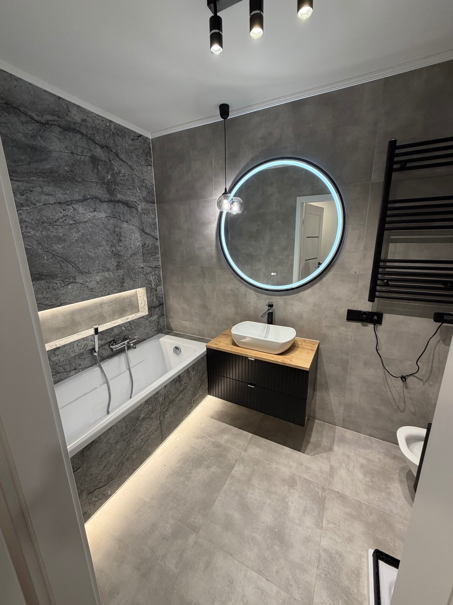 Nowoczesna łazienka w szarościach z podświetlaną wanną, okrągłym lustrem LED i drewnianą szafką pod umywalką. Minimalistyczny design z czarnym grzejnikiem.