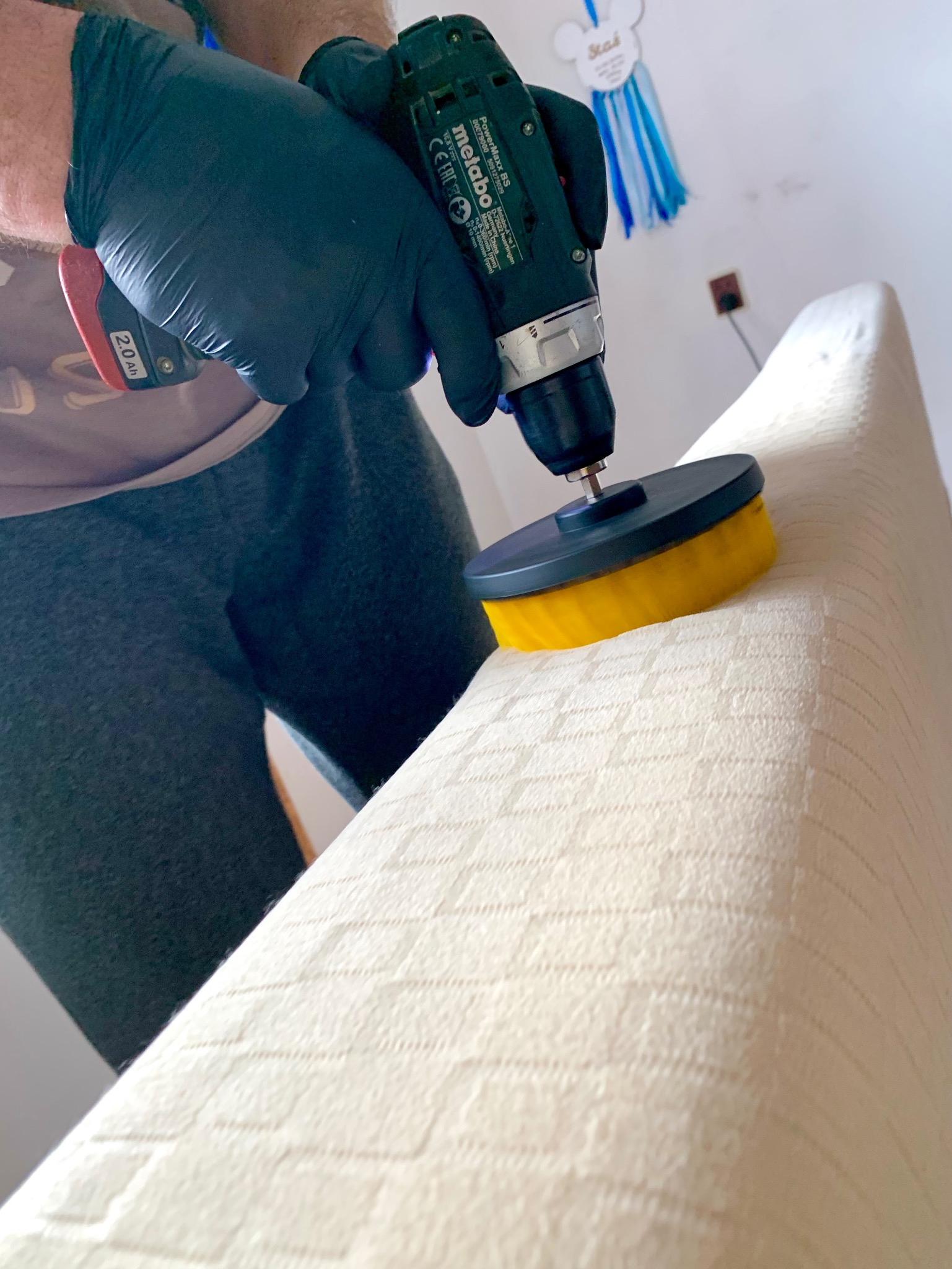 Czyszczenie jasnej tapicerki kanapy w Rybniku przy użyciu wiertarki z okrągłą szczotką przez osobę w czarnych rękawicach ochronnych.