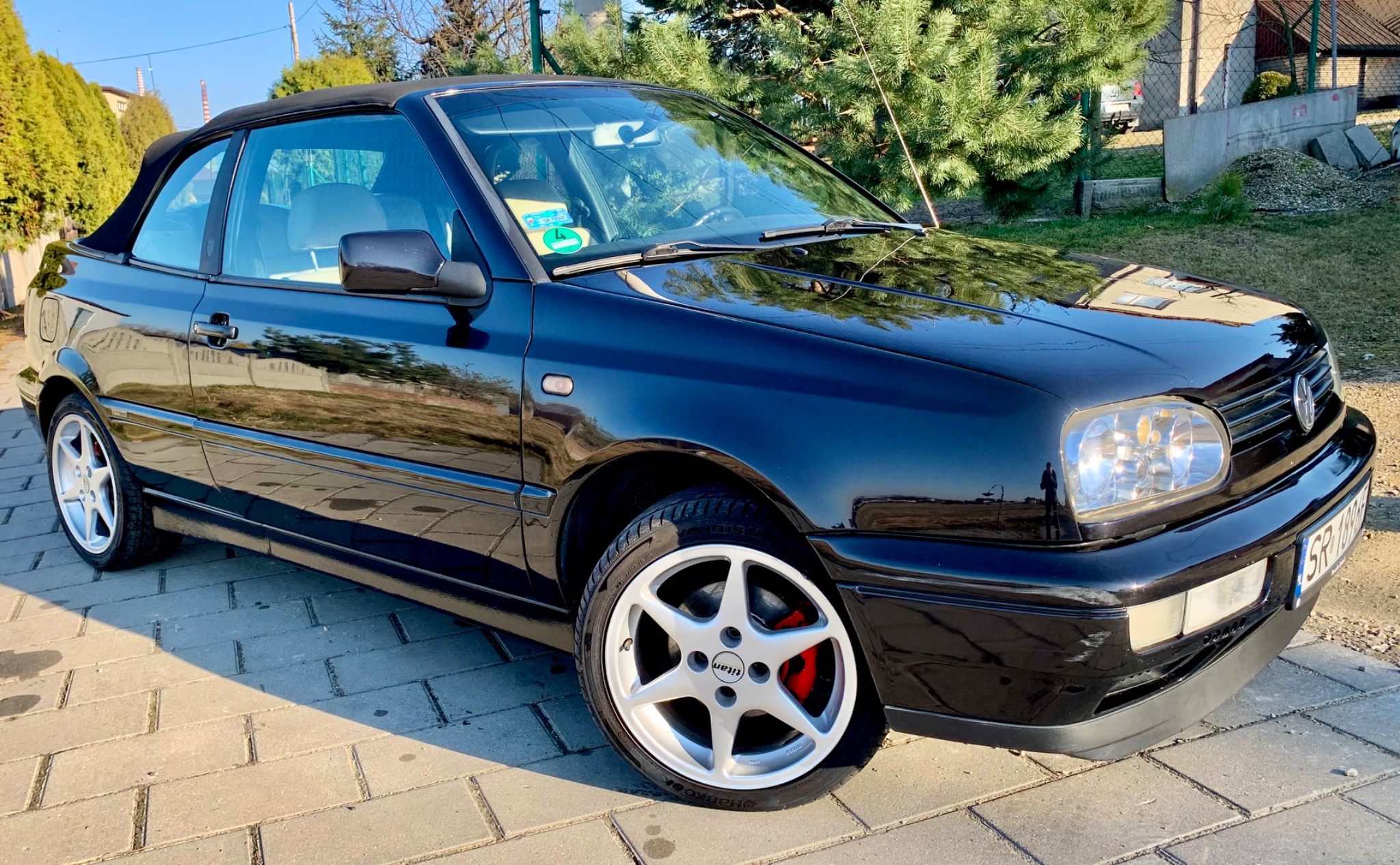Błyszczący, czarny kabriolet Volkswagen Golf III na kostce brukowej, z widocznymi srebrnymi felgami Titan i czerwonymi zaciskami hamulcowymi, w tle zielone drzewa i budynek.
