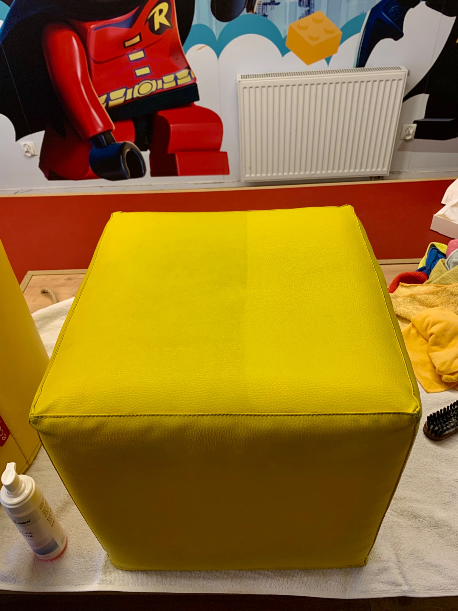 Żółty puf z ekoskóry, widoczna różnica w czystości połowy powierzchni po zabiegu czyszczenia, w tle ściana z motywem Lego, obok butelka środka czyszczącego i szczotka