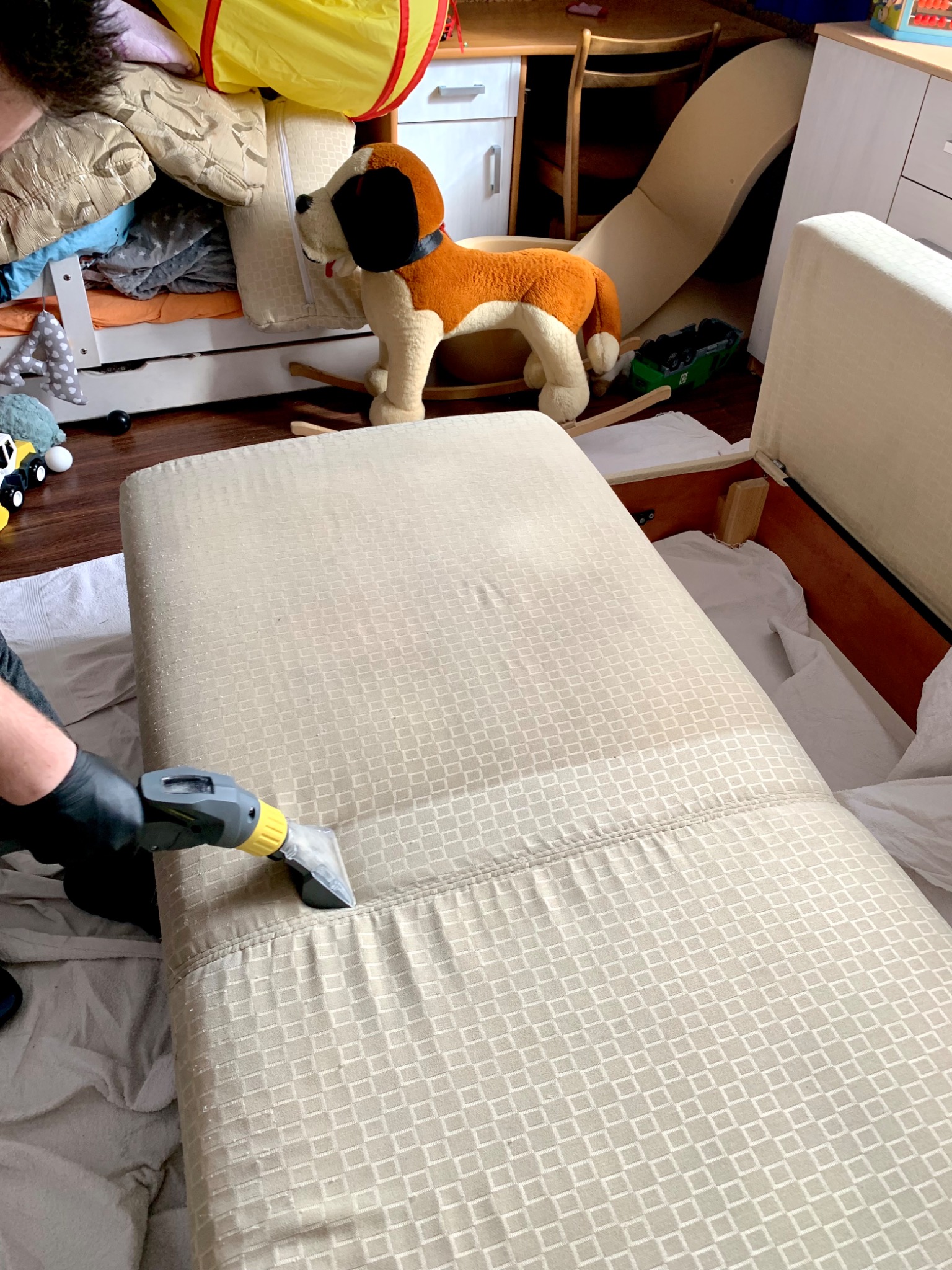 Czyszczenie tapicerki kanapy z wzorem w kwadraty za pomocą odkurzacza piorącego, widoczne efekty czyszczenia i otwarte skrzynie kanapy w pokoju dziecięcym.