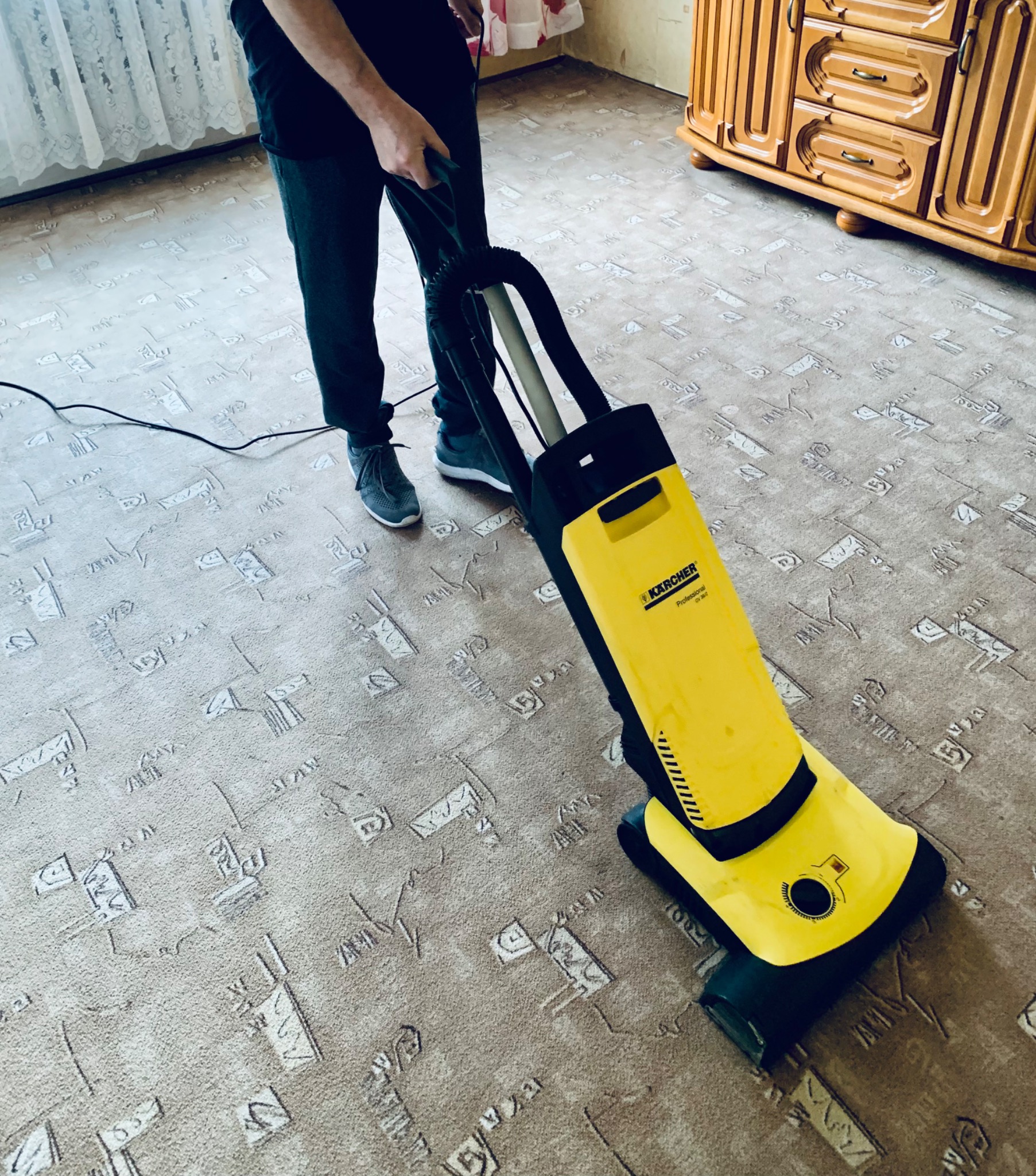 Osoba w ciemnych spodniach i szarych butach obsługuje żółto-czarny odkurzacz Kärcher Professional CV 38/1 na beżowym dywanie we wzory, obok stoi drewniana komoda.