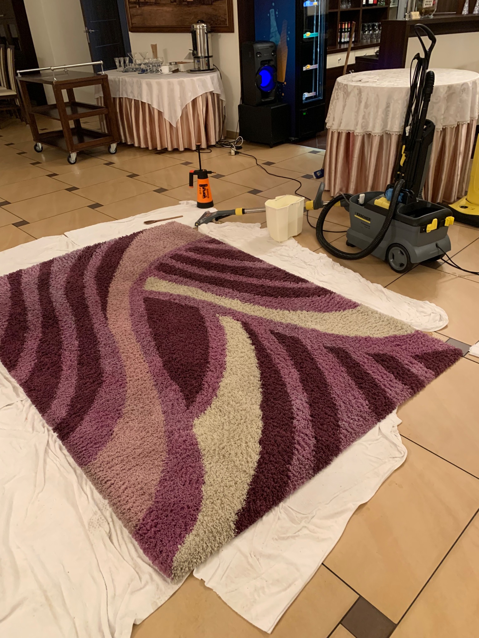 Wzorzysty, fioletowo-beżowy dywan leżący na białym prześcieradle, obok spryskiwacz ciśnieniowy, odkurzacz piorący Kärcher i wiadro, w tle stoliki nakryte białymi obrusami.