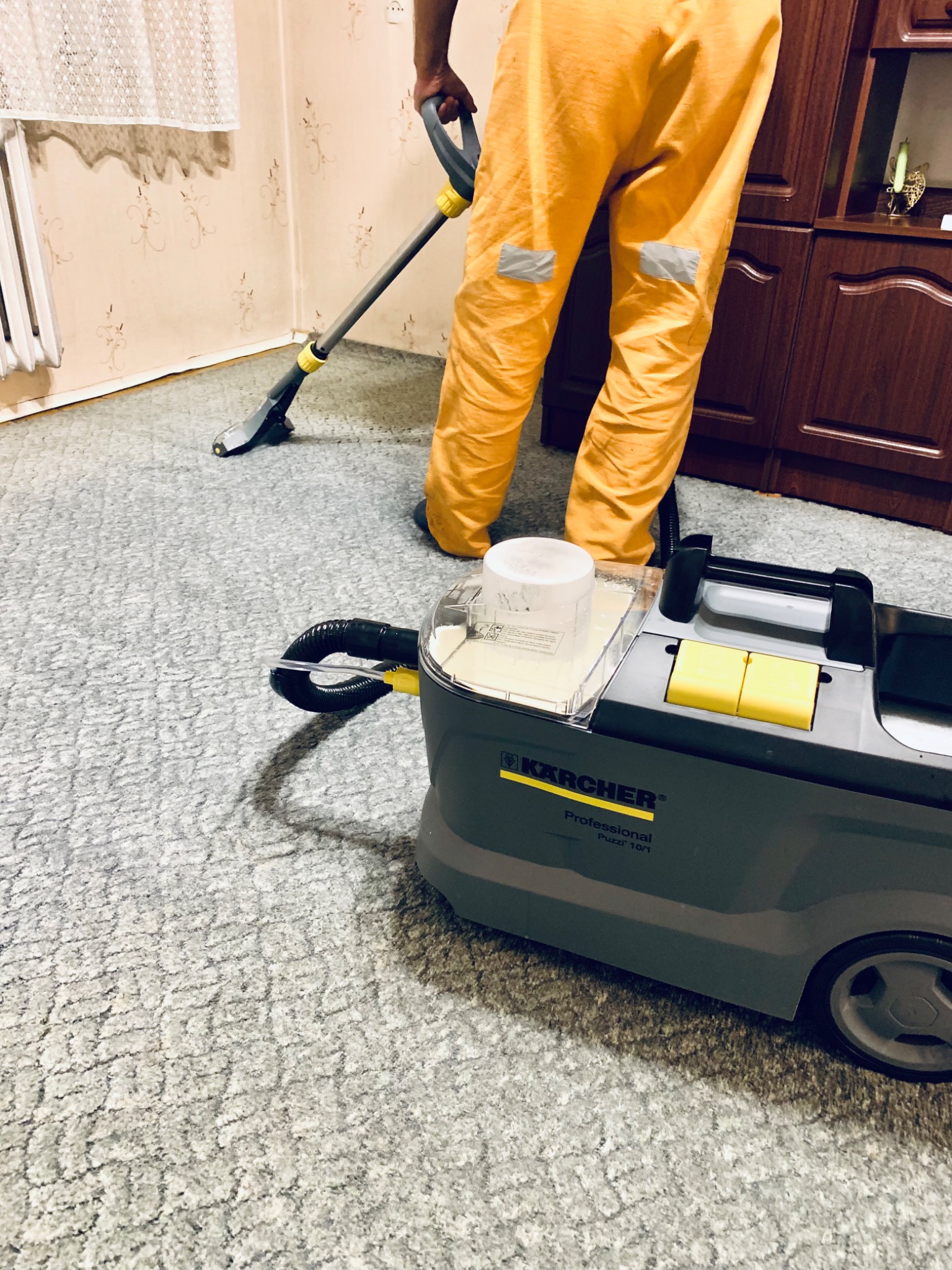 Osoba w pomarańczowych spodniach roboczych czyści jasnoszary dywan urządzeniem Kärcher Puzzi 10/1, widoczne ślady wilgoci po czyszczeniu.