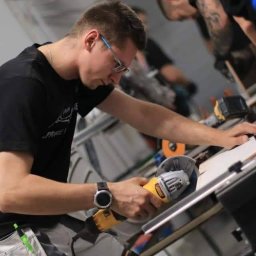 Pogotowie remontowe michał lewandowski - Mężczyzna w okularach tnie płytkę ceramiczną szlifierką kątową na stole warsztatowym, w tle widoczni inni pracownicy.