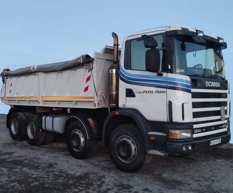 Ciężarówka Scania, biała kabina z niebieskimi pasami, wywrotka z plandeką, widoczne oznaczenia ostrzegawcze na burcie, firma Pavel-Trans.