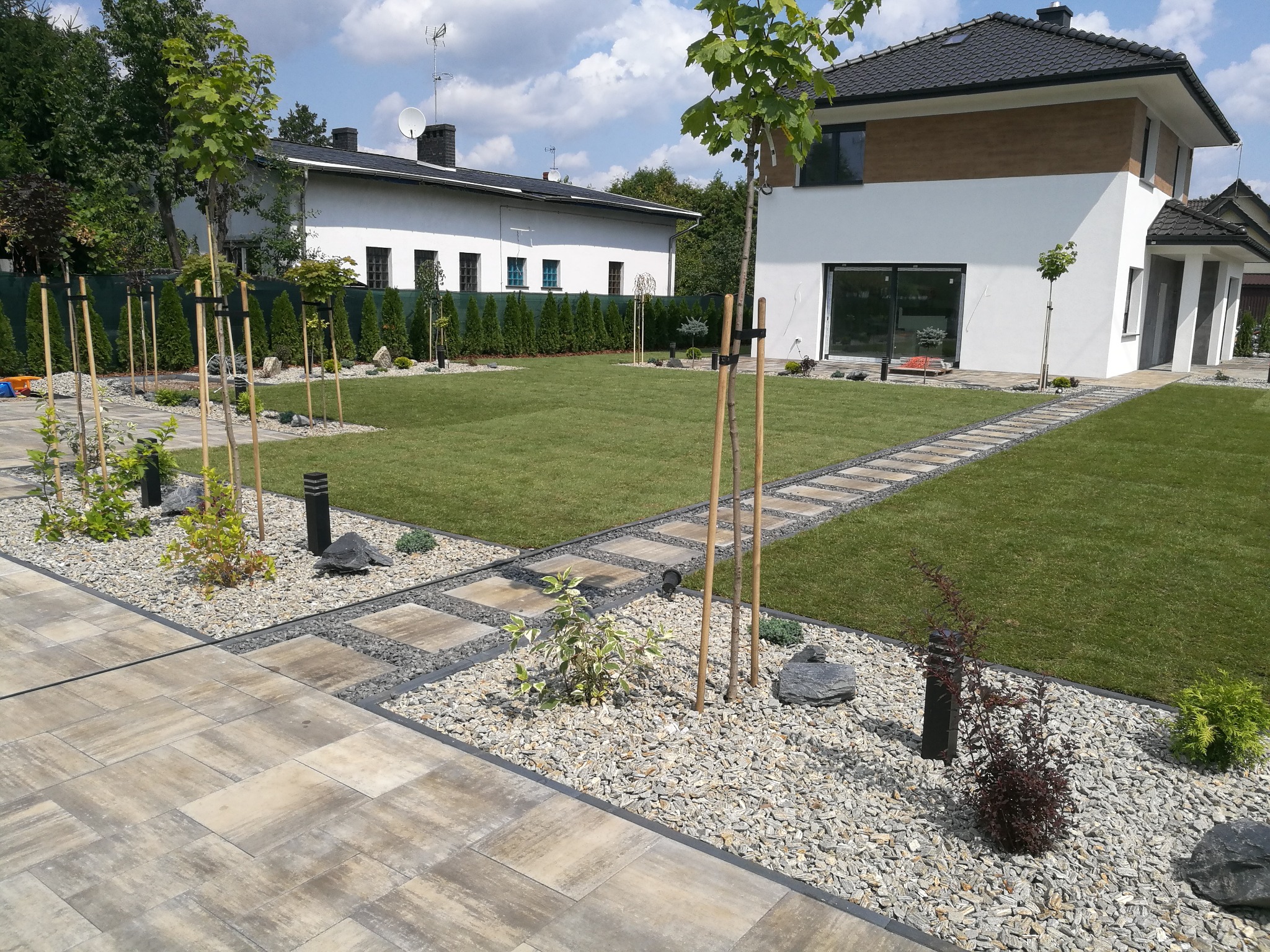 Nowoczesny ogród z równo przystrzyżonym trawnikiem, ścieżką z płyt betonowych, rabatami z kamienia i młodymi drzewkami podpartymi tyczkami.