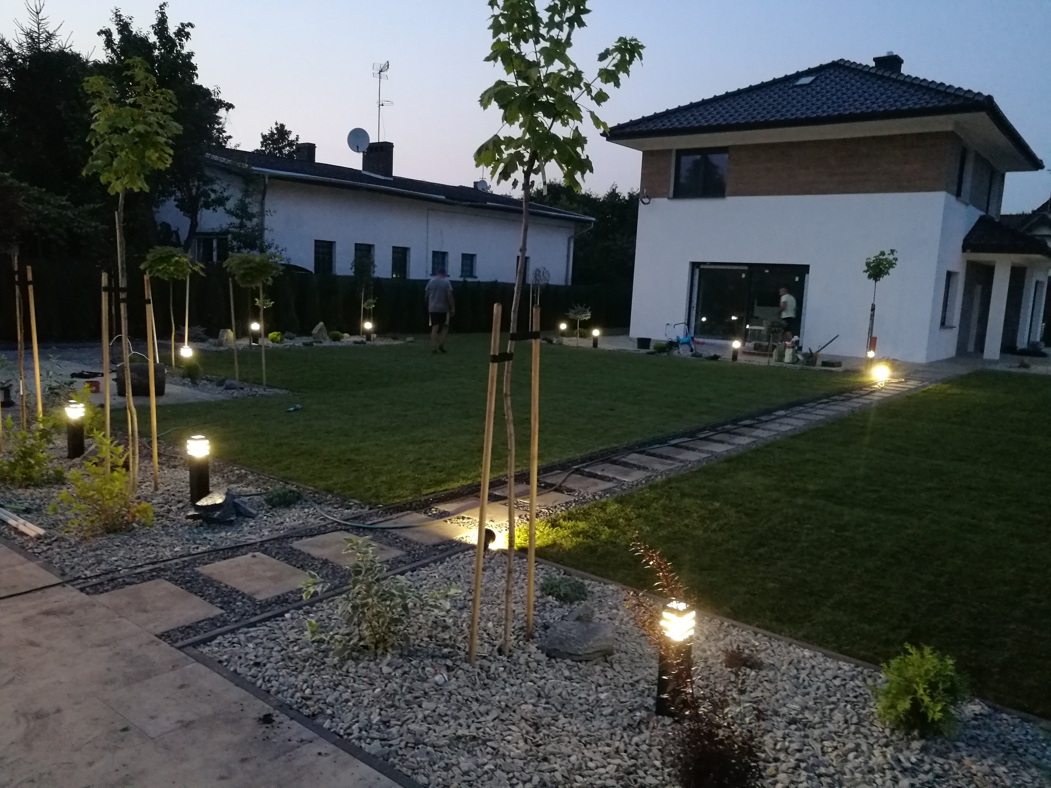 Oświetlony ogród po zmierzchu z młodymi drzewkami, ścieżką z płyt i żwiru, oraz lampami ogrodowymi, na tle nowoczesnego domu.
