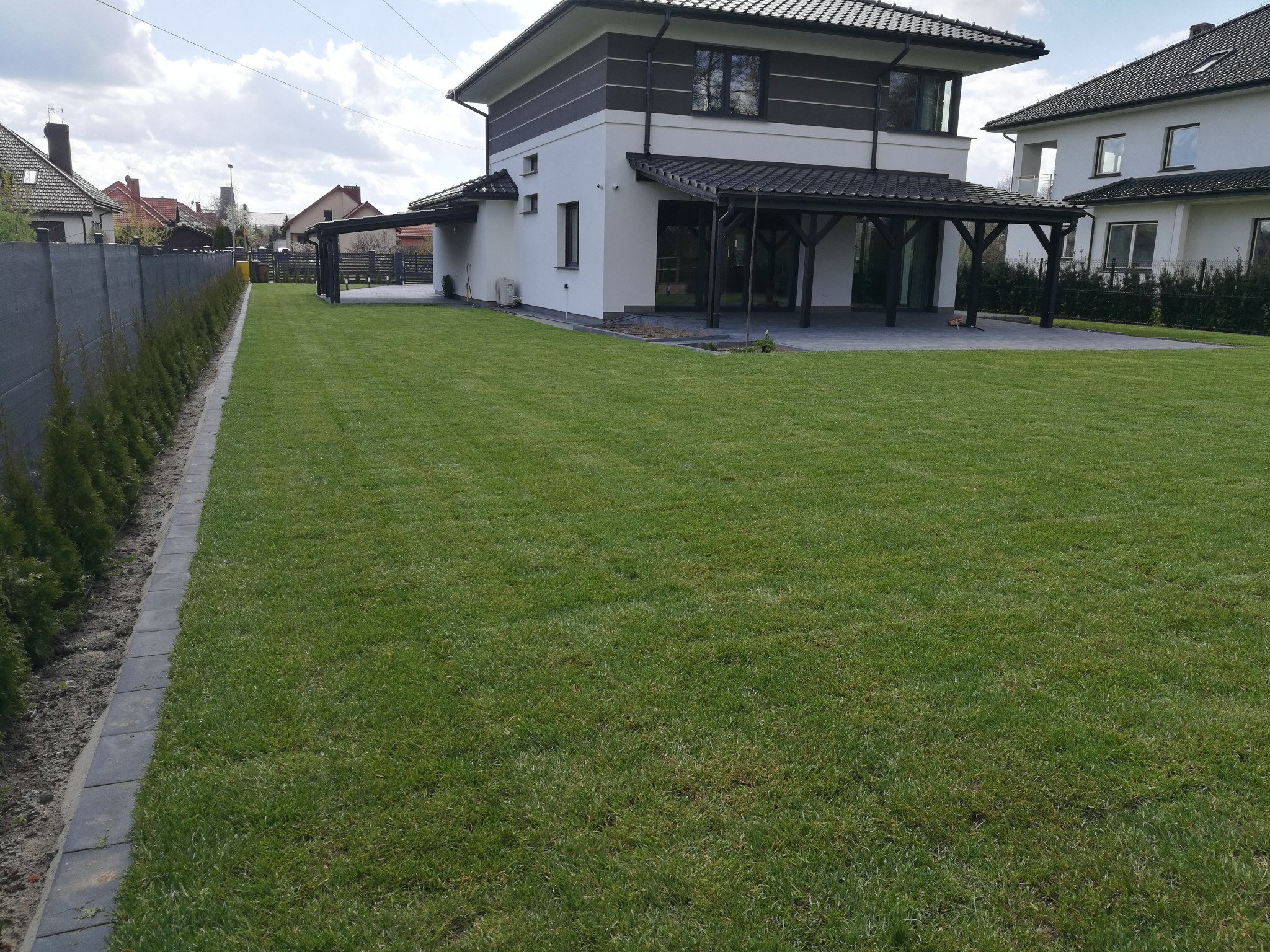 Starannie utrzymany trawnik przed nowoczesnym domem z szarą elewacją i zadaszeniem tarasu, obrzeża z szarych kostek i rząd małych tuj wzdłuż betonowego ogrodzenia.