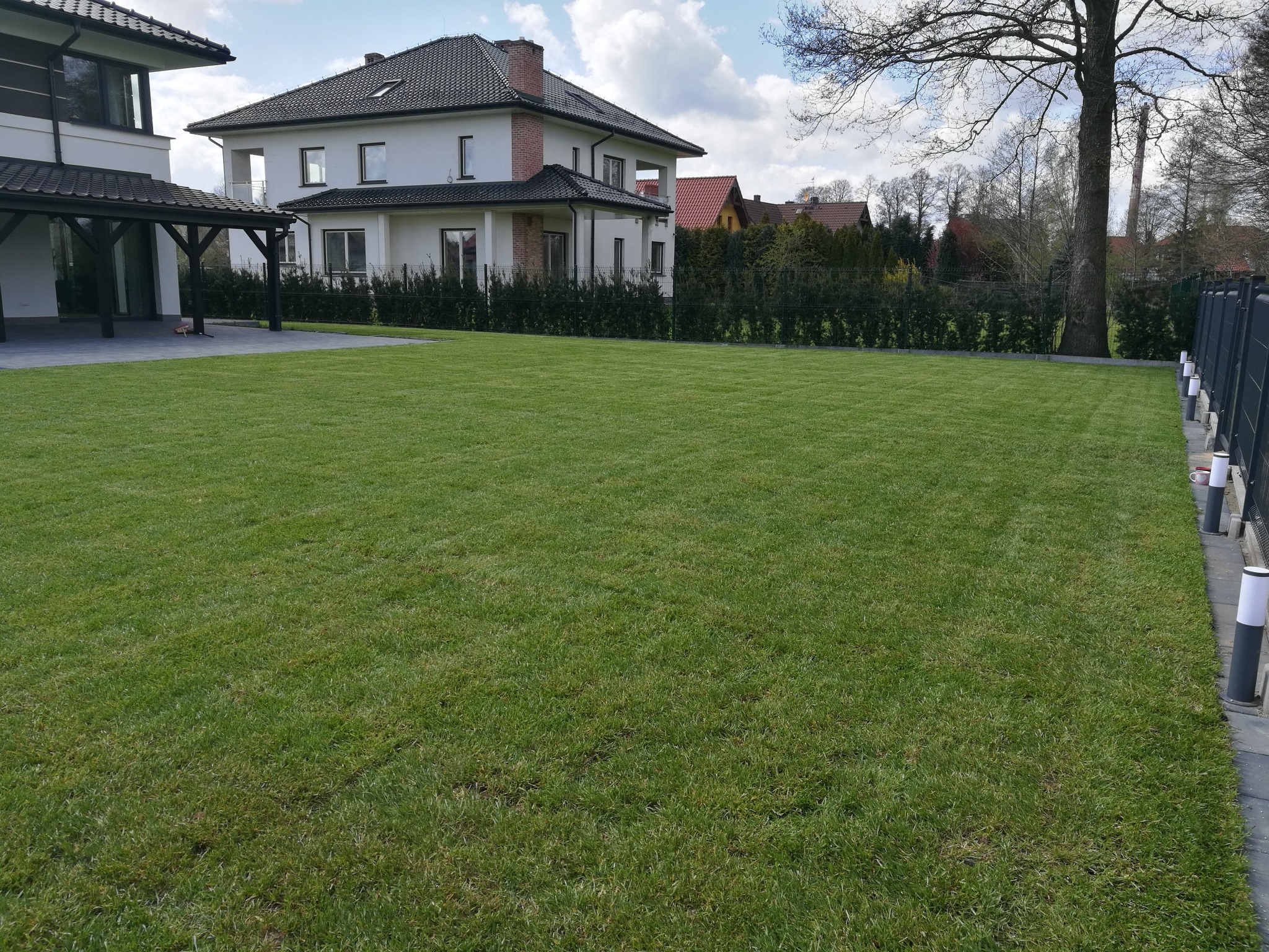 Rozległy, równo skoszony trawnik przed nowoczesnym domem z ciemną dachówką i ogrodzeniem z lampami, widok z perspektywy trawy.