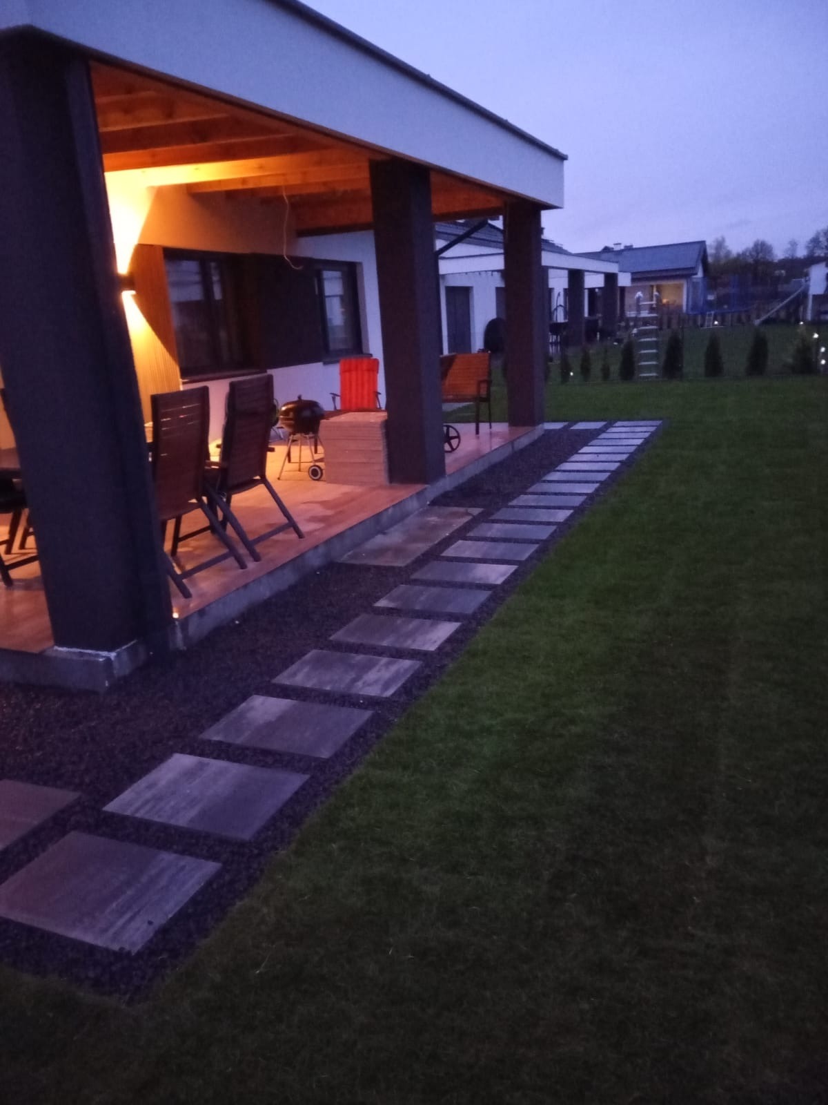 Nowoczesny ogród z równo przystrzyżonym trawnikiem, ścieżką z kwadratowych płyt i zadaszonym tarasem z grillem, oświetlonym ciepłym światłem.