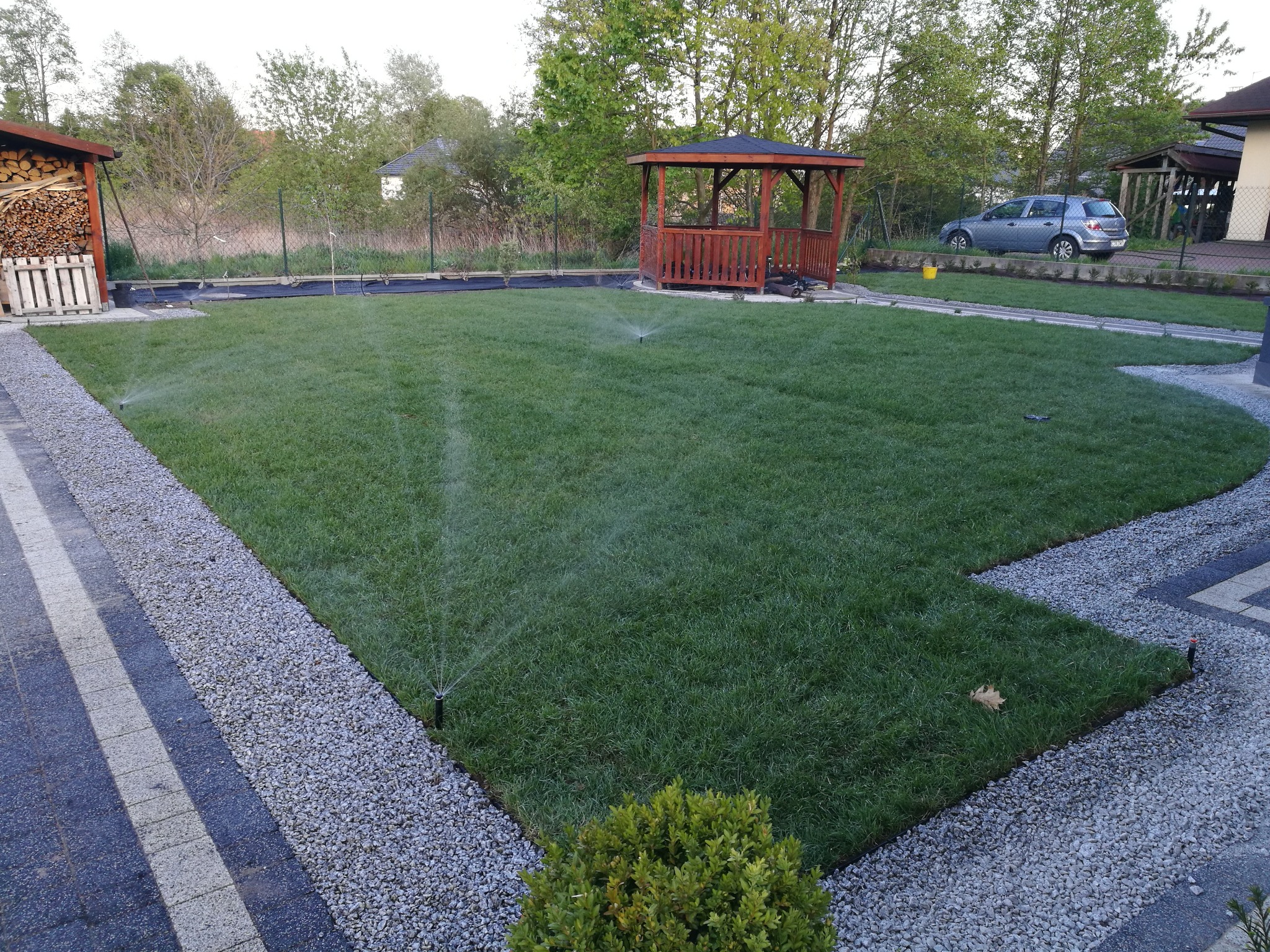 Nawadniany trawnik z drewnianą altaną w tle, obrzeża z jasnych kamyków i kostki brukowej, widoczne zraszacze.