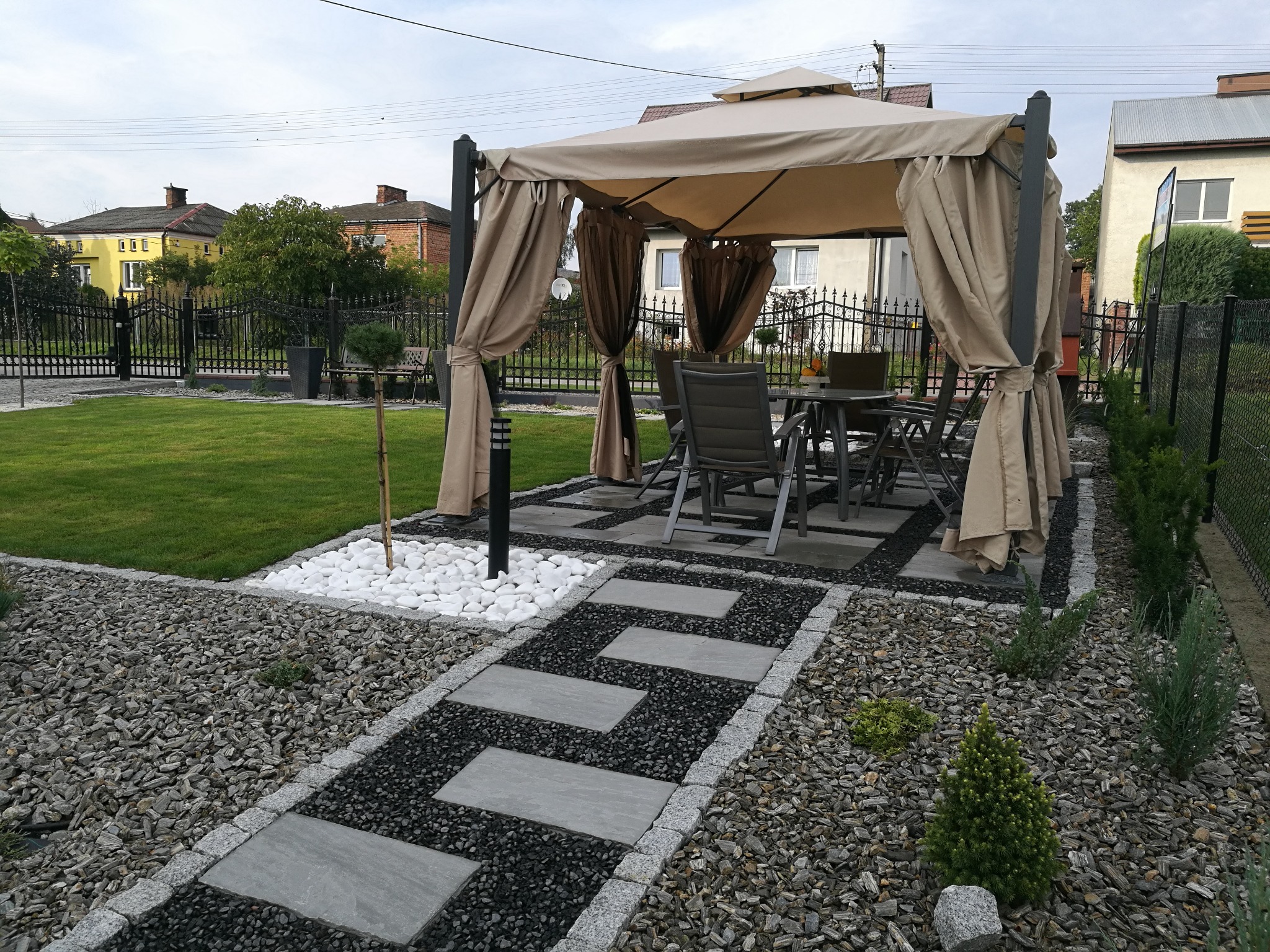Ogród z beżową altaną, szarymi meblami ogrodowymi, ścieżką z płyt i białymi kamieniami dekoracyjnymi.