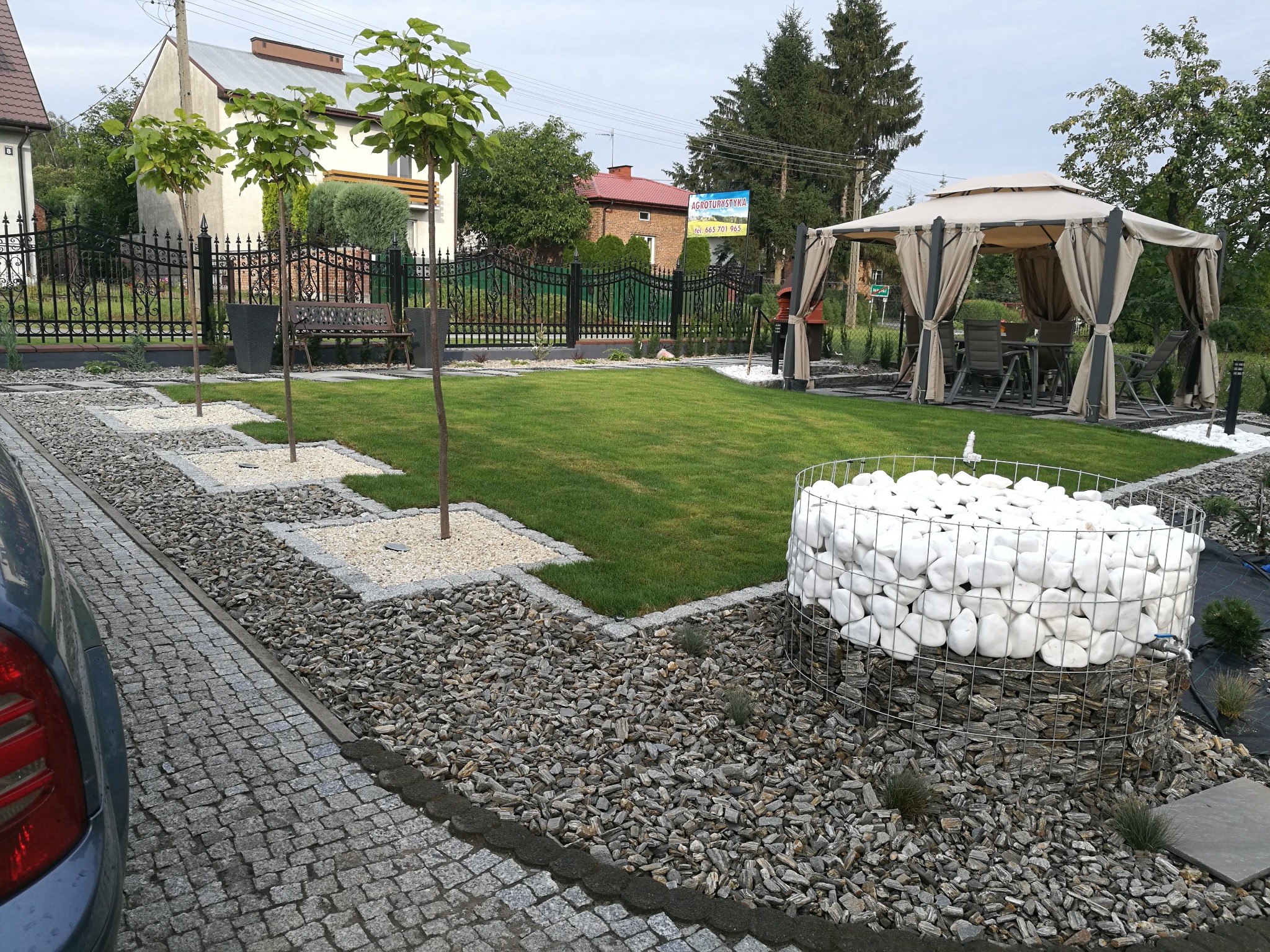 Zadbany ogród z równo przystrzyżonym trawnikiem, ozdobnymi drzewkami w kwadratowych rabatach z białym żwirem, gabionem wypełnionym białymi kamieniami i altaną ogrodową w tle.