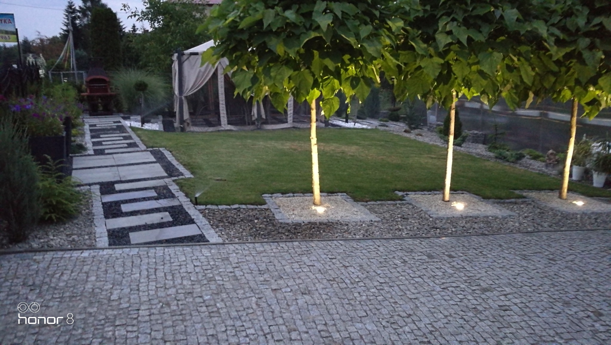 Oświetlony ogród z kostki brukowej, ścieżki z płyt i żwiru, trawnika oraz drzew z podświetleniem u podstawy, widoczny namiot ogrodowy w tle.