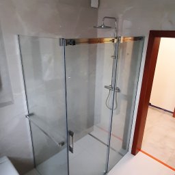 LukFed - Szklana kabina prysznicowa z chromowanymi okuciami w jasnej łazience z marmurowymi płytkami, widoczny fragment toalety i drzwi do innego pomieszczenia z pomarańczowym zabezpieczeniem podłogi.