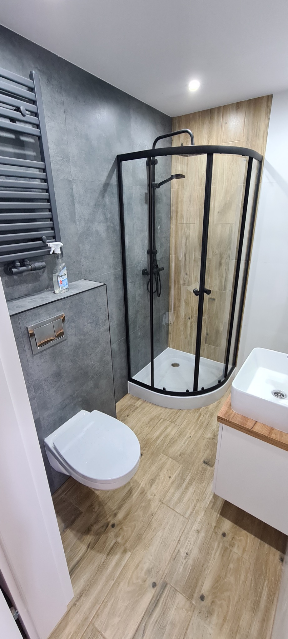 Nowoczesna łazienka po renowacji: kabina prysznicowa z czarnymi ramami, wisząca toaleta, umywalka nablatowa na drewnianej szafce, szare płytki ścienne i podłogowe imitujące drewno, grzejnik...
