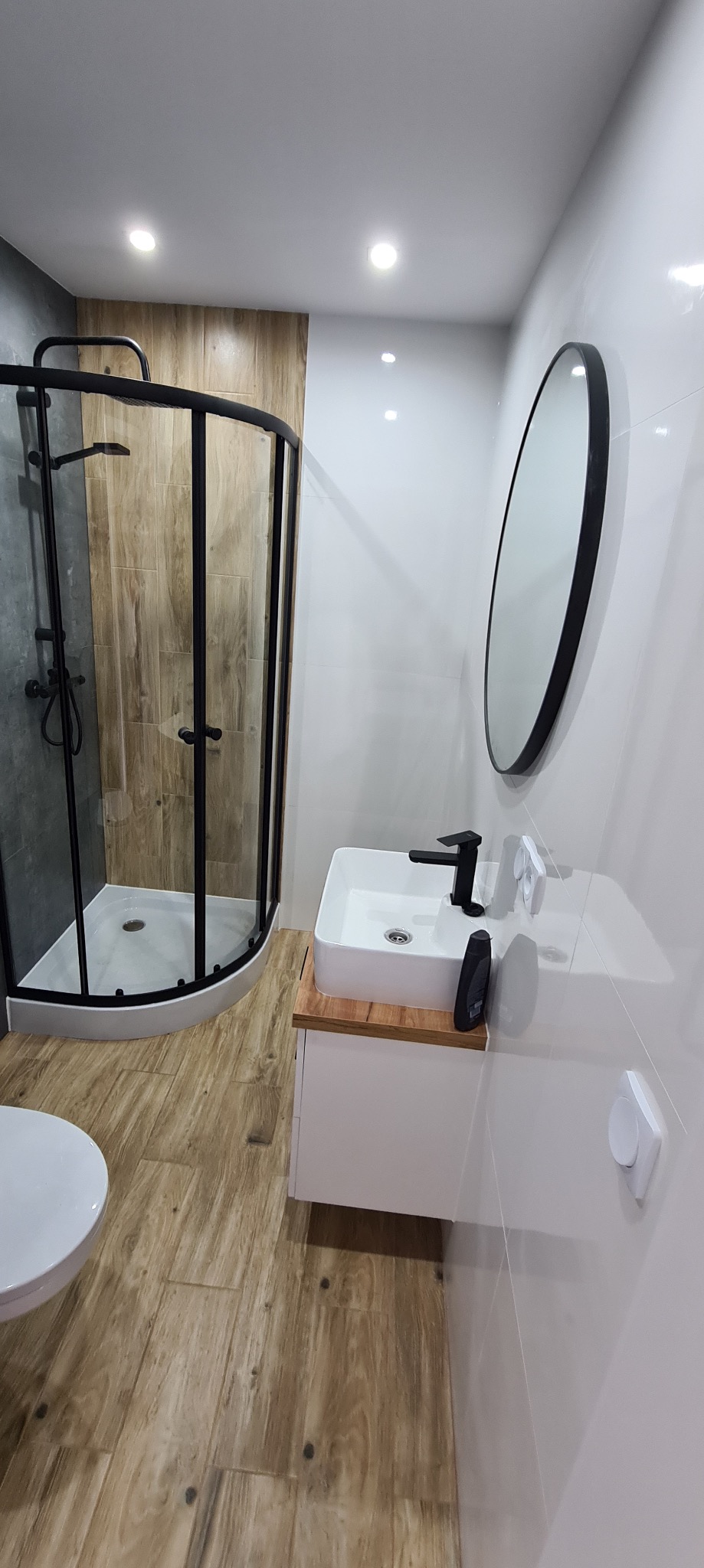 Nowoczesna łazienka z kabiną prysznicową typu walk-in z czarnymi profilami, owalnym lustrem w czarnej ramie, białą umywalką nablatową z czarną armaturą i płytkami imitującymi drewno na podłodze...